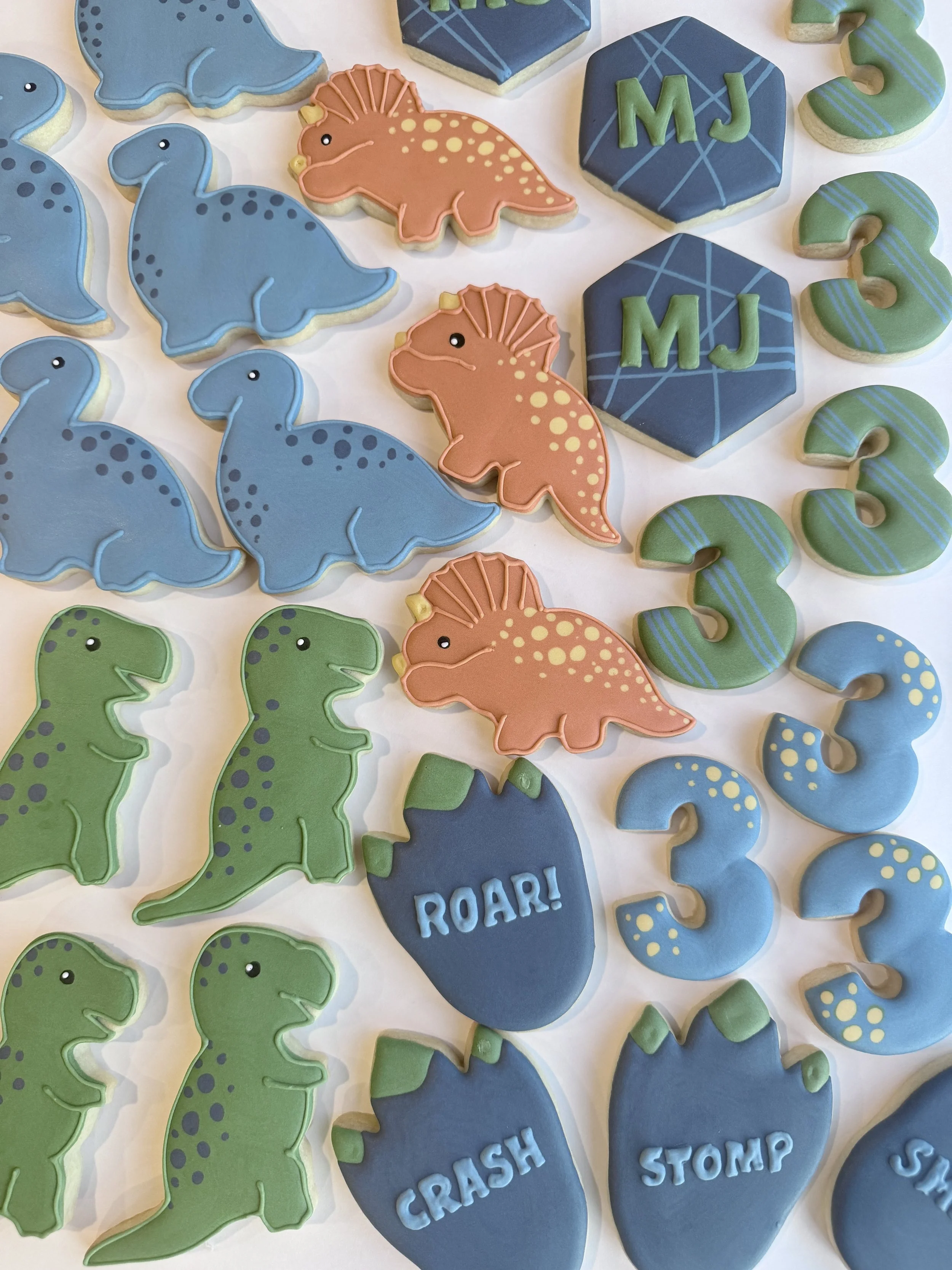 A collection of dinosaur-themed cookies decorated with colored icing, including blue dinosaurs, orange brachiosaurus, and green dinosaurs, along with blue and green number cookies, and blue cookies with words like 'ROAR!', 'CRASH', and 'STOMP' writte