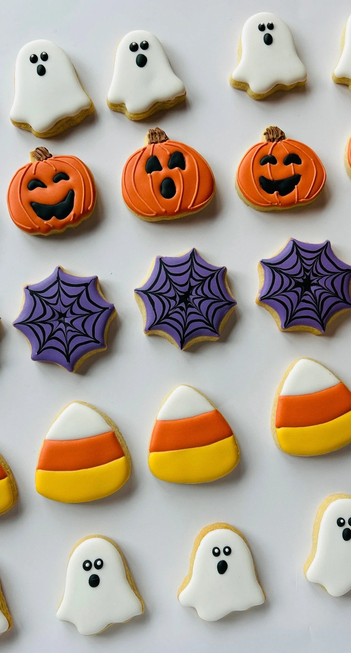 Halloween themed decorated cookies featuring white ghost shapes, orange jack-o'-lanterns, purple spiderwebs, and candy corn with white, orange, and yellow icing.