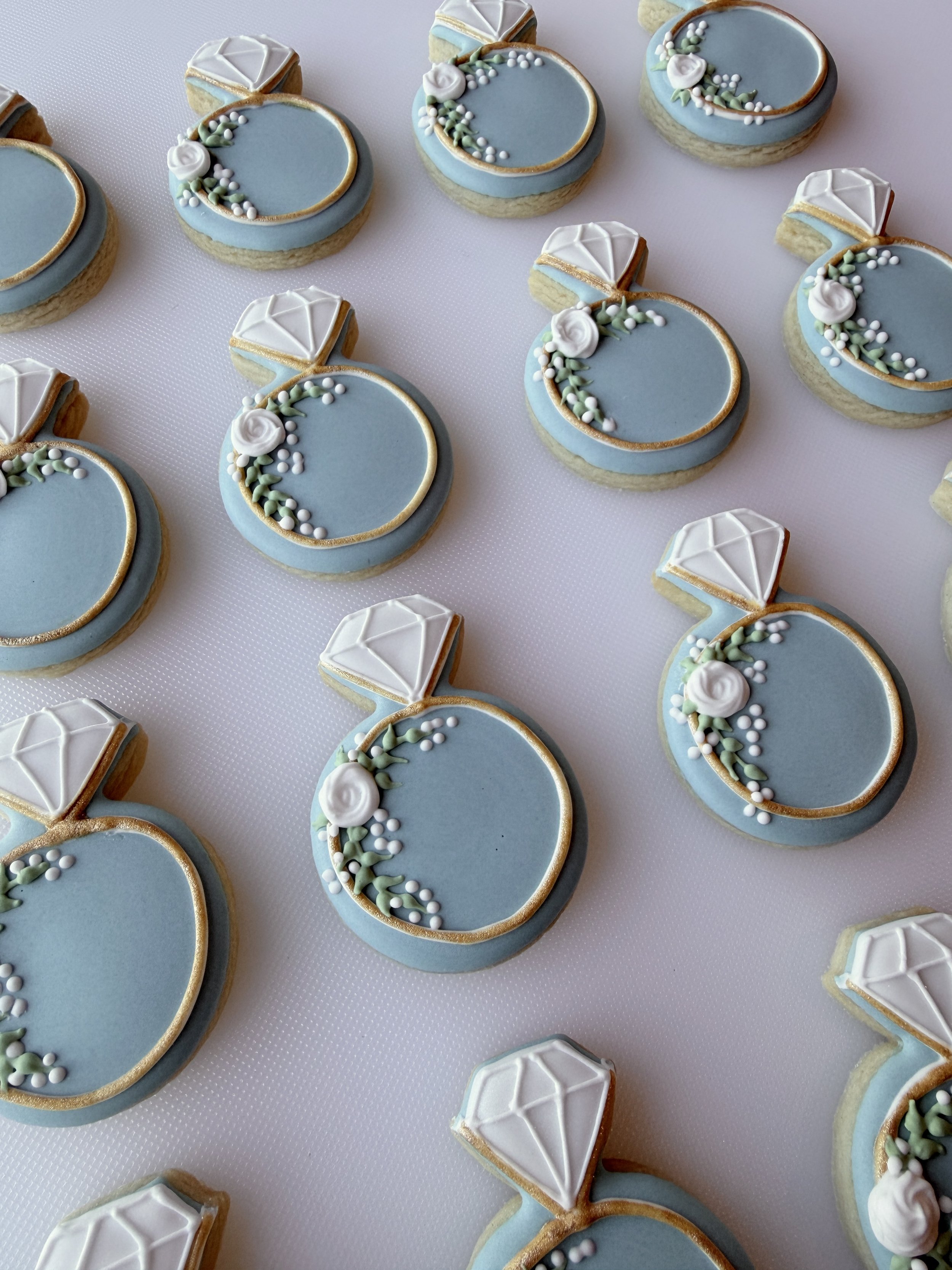 A variety of cookies decorated with blue icing, white roses, green leaves, and small white and green sugar balls, some shaped like diamonds.