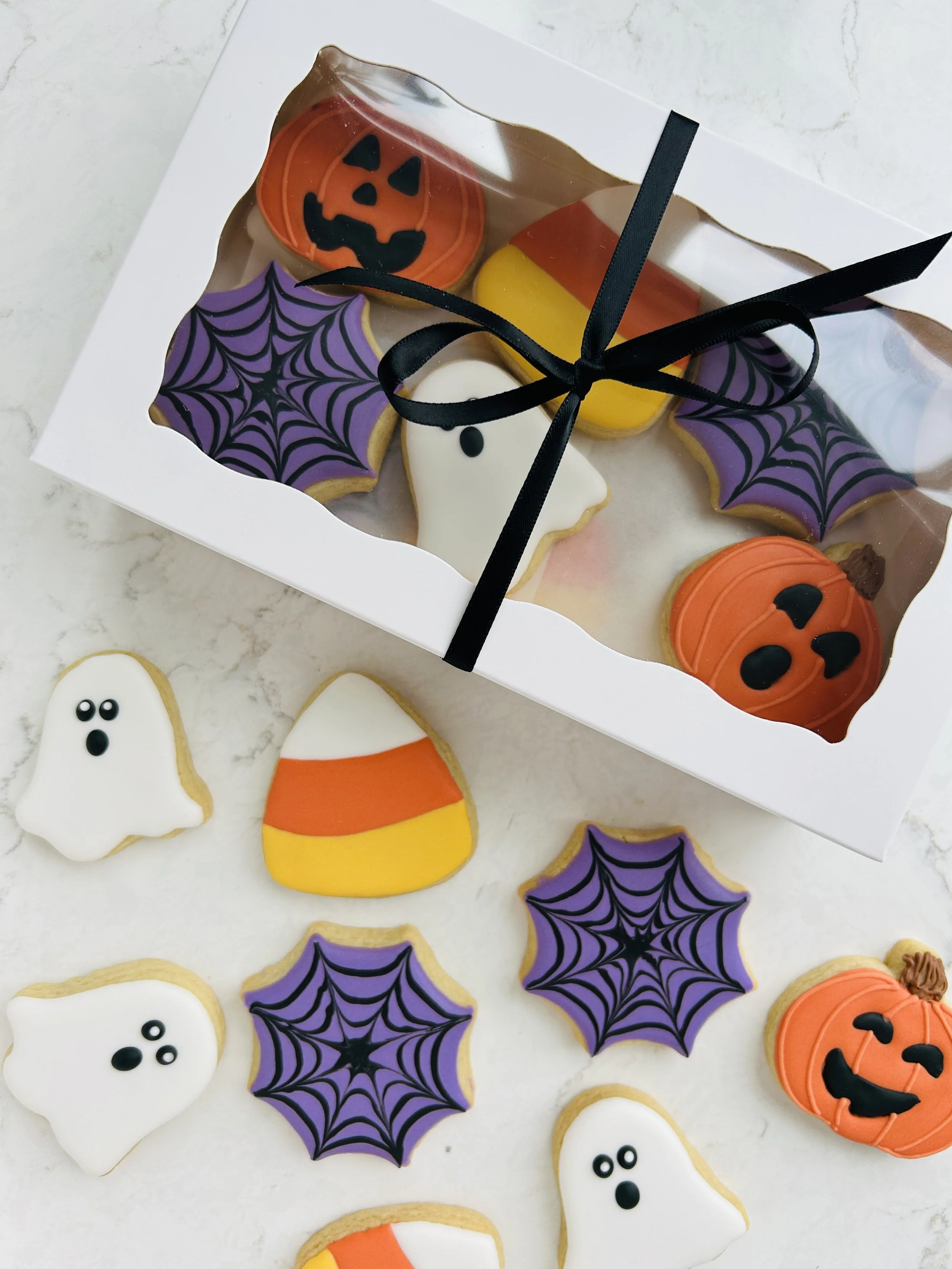 Assorted Halloween-themed cookies including ghost shapes with black eyes, orange pumpkin shapes with black faces, purple bat shapes with black spiderweb patterns, and candy corn shapes with orange, yellow, and white sections, inside a clear plastic g