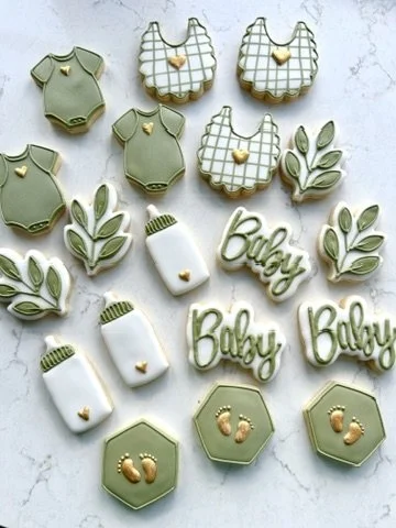 Baby-themed cookies including onesies, bibs, bottles, leaves, footprints, the word 'Baby', and paw prints, decorated in pastel green, white, and gold accents.