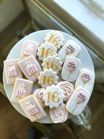 A round birthday cake decorated with pink and white themed treats, including a large number 16, candles, and birthday message cookies, all on a white plate.