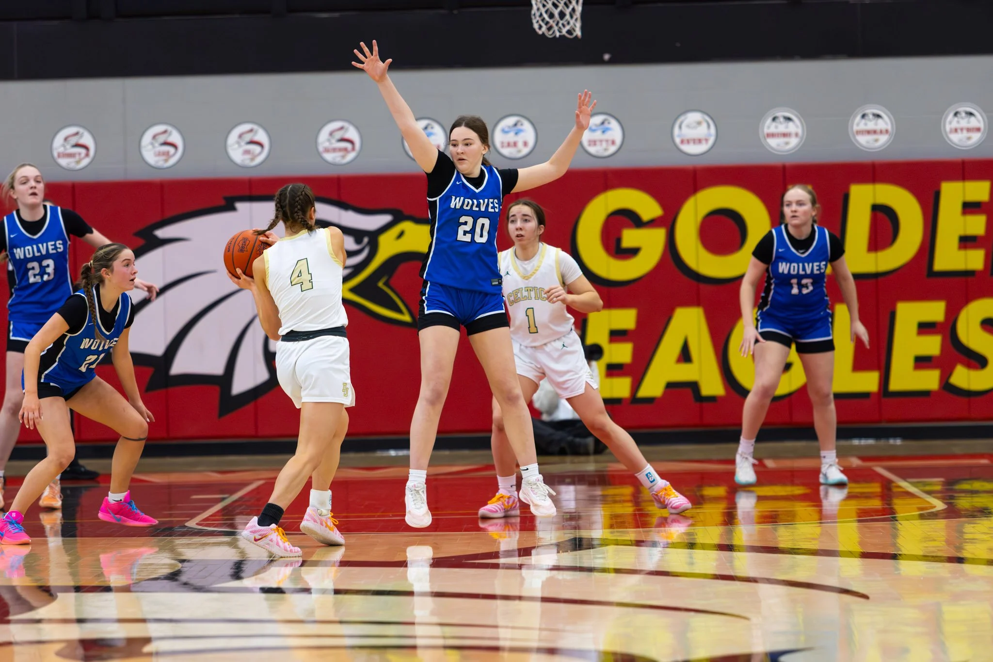 Girls playing basketball during a game, with one girl in white holding the ball and others in blue defending, on a gymnasium court with a large logo and red wall in the background.