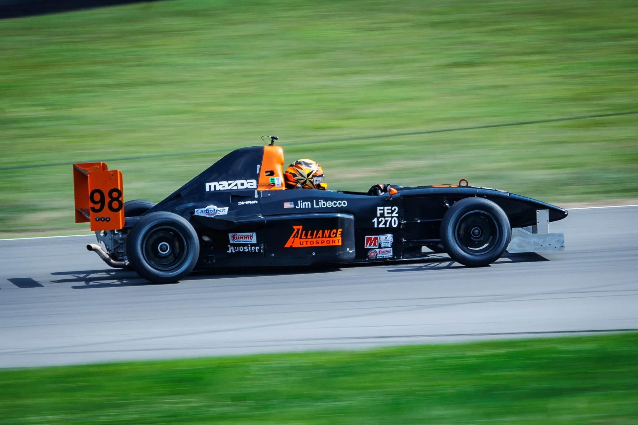 Black race car with orange accents speeding on a track, labeled with sponsor logos and the number 98, driven by Jim Libecco.