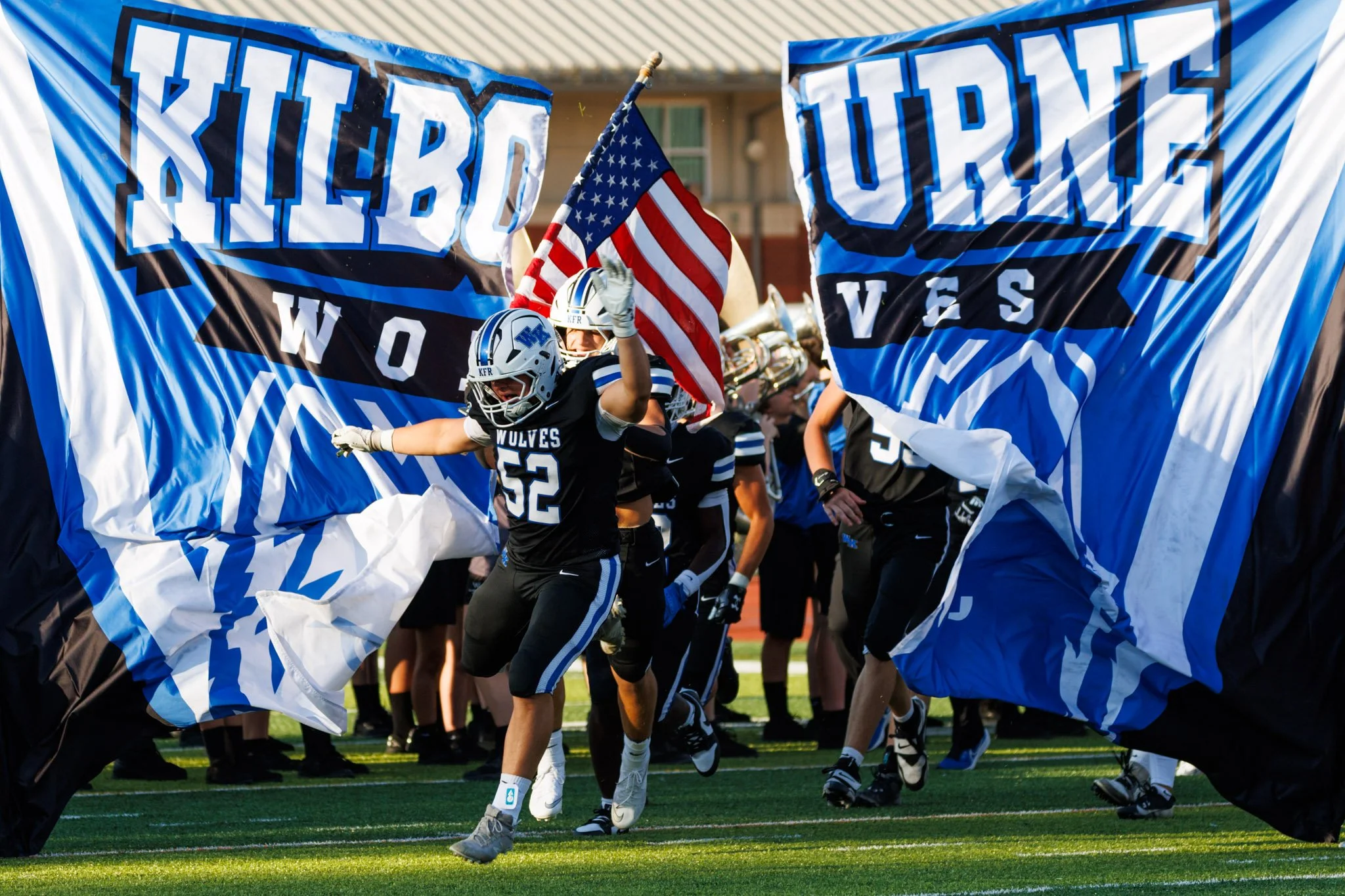 High school football players in black and blue uniforms running onto the field through large banners that say 'Kilbourne Wolves' as they run onto the field with an American flag.