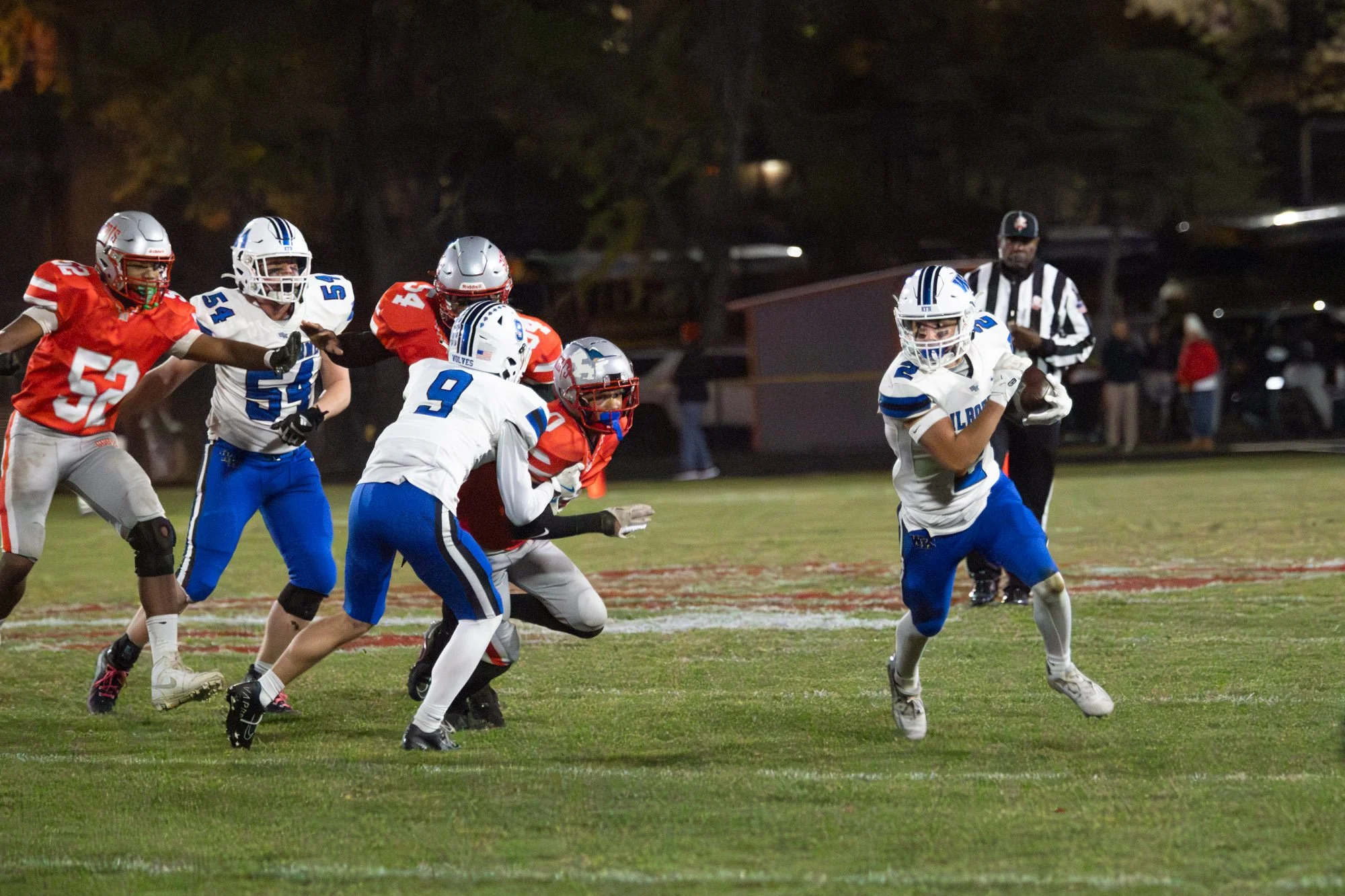 A football game at night with players in red and white uniforms. A player in a white and blue uniform is running with the football, pursued by a player in red attempting to tackle him. Several other players and a referee are visible in the background