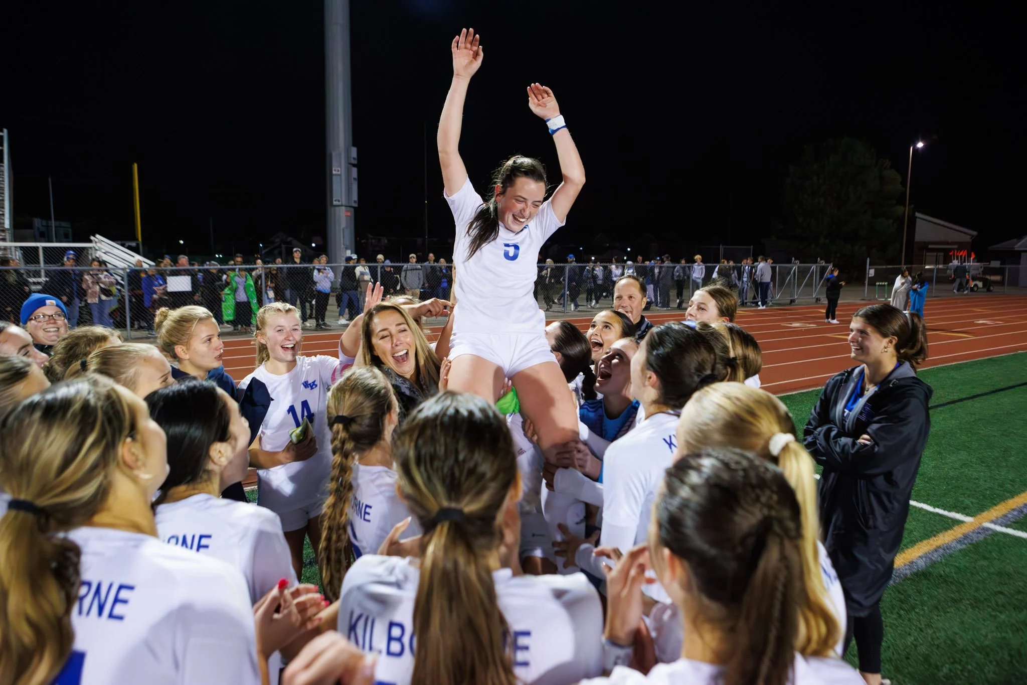 A female soccer player being lifted onto her teammates' shoulders during a celebration on a soccer field at night.