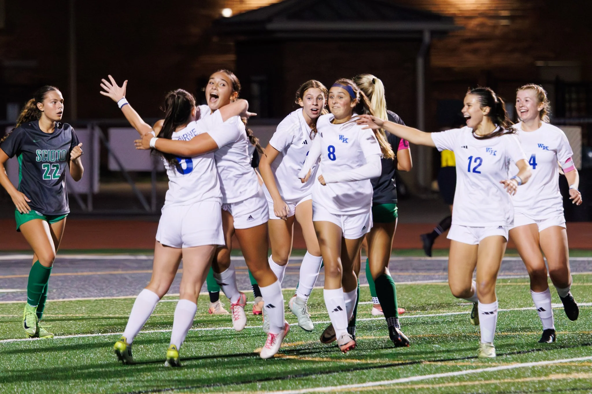 Girls soccer team celebrating on the field at night, wearing white jerseys and shorts, some with numbers 8, 12, 4, and others, with one girl in black and green uniform.