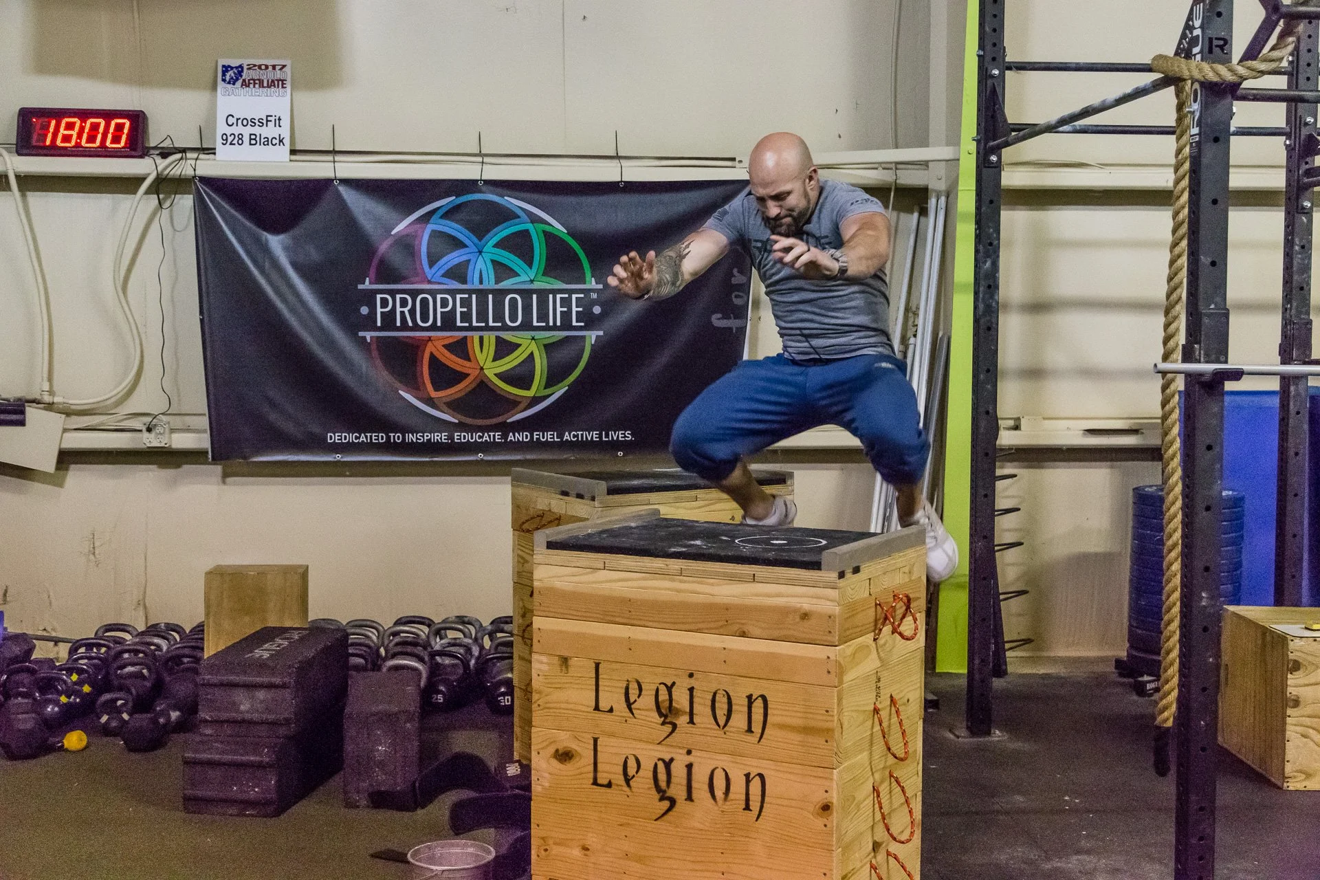 A man is jumping onto a wooden plyometric box at a gym, with kettlebells and other equipment nearby. There is a CrossFit Propello Life banner in the background and a digital clock on the wall.