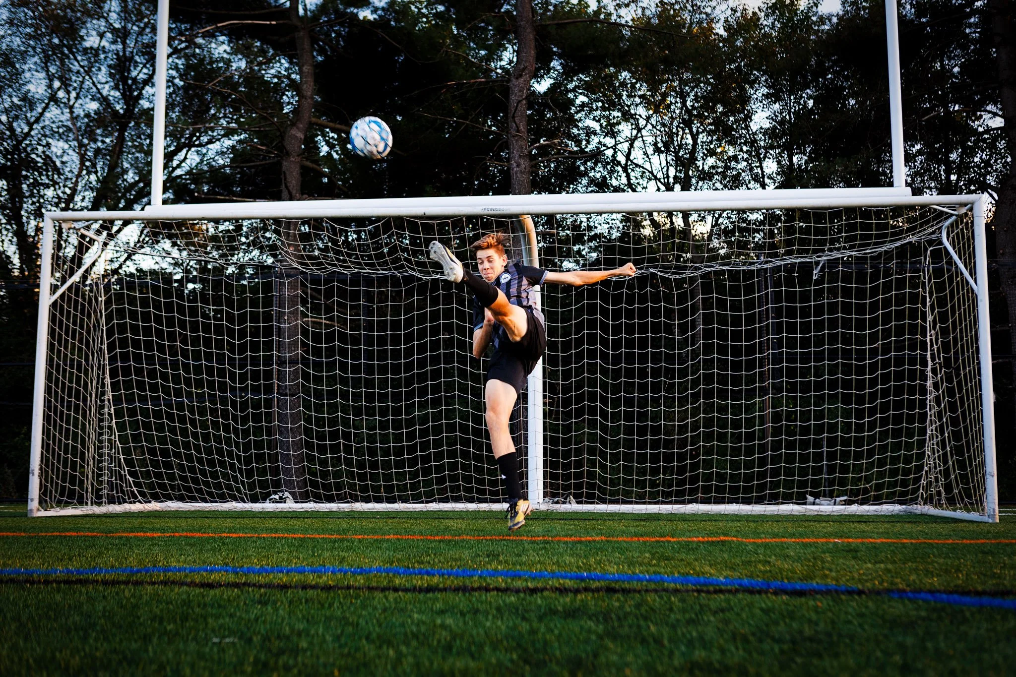 A soccer goalkeeper in black shorts and a black jersey with a gray stripe, jumping in the air to make a save during a game, with a soccer ball in midair near the goal.