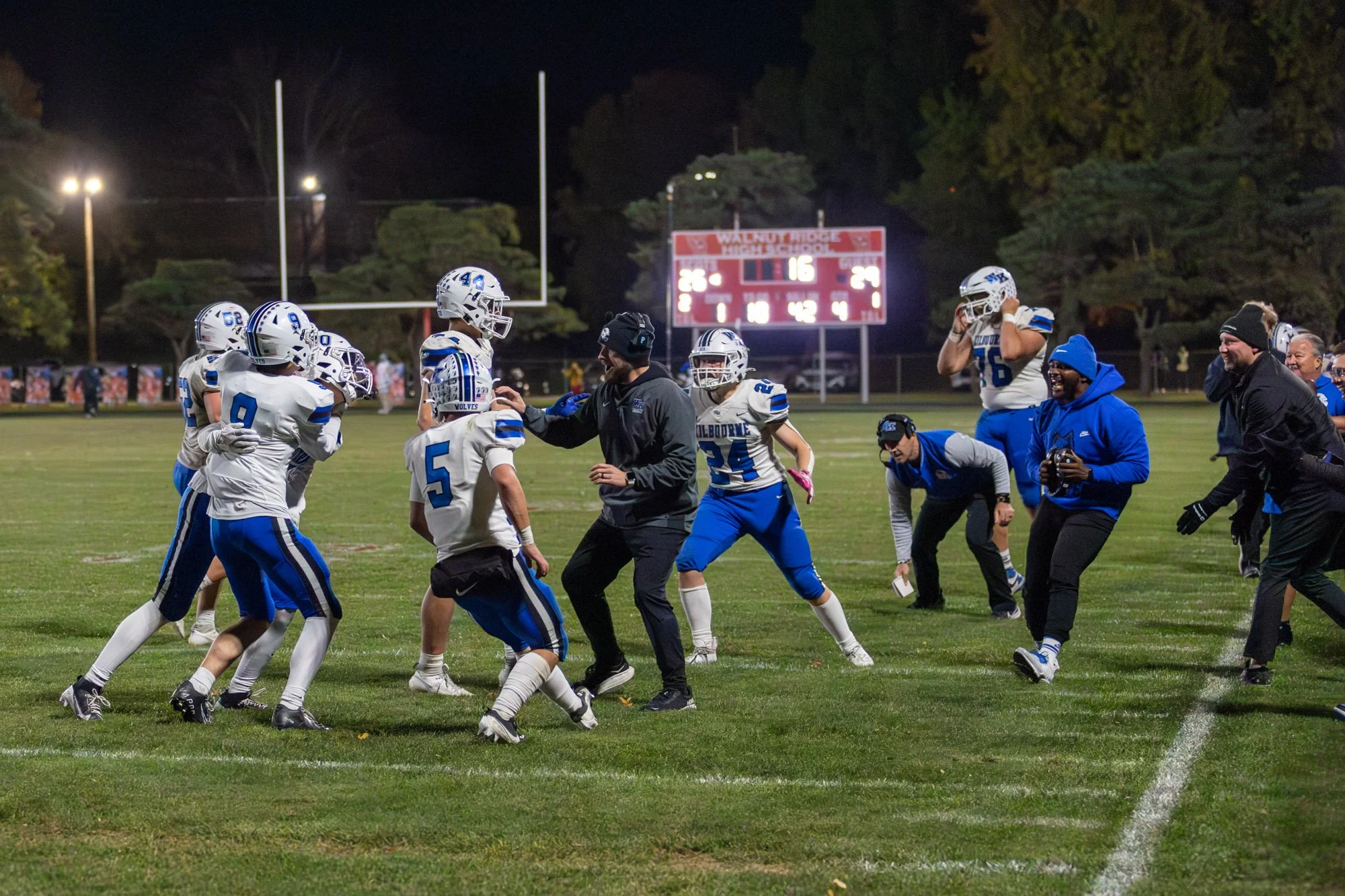 Nighttime football game on a grassy field with players wearing blue and white uniforms and a scoreboard in the background.
