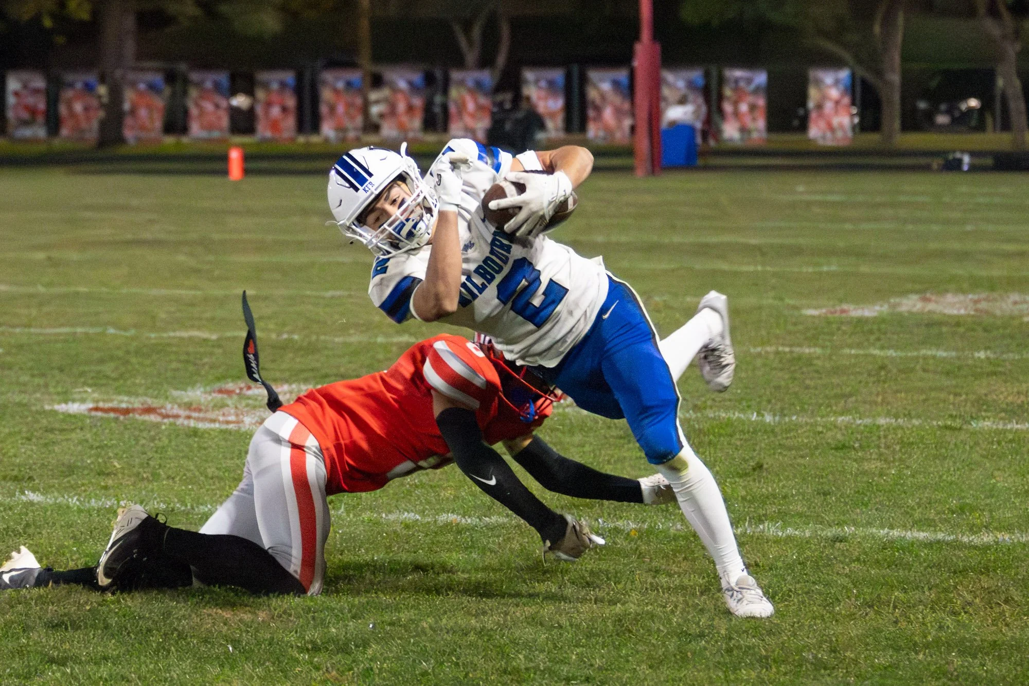 A football player in a white and blue uniform with the number 12 is running with the ball while an opponent in red and white attempts to tackle him on a grassy field.