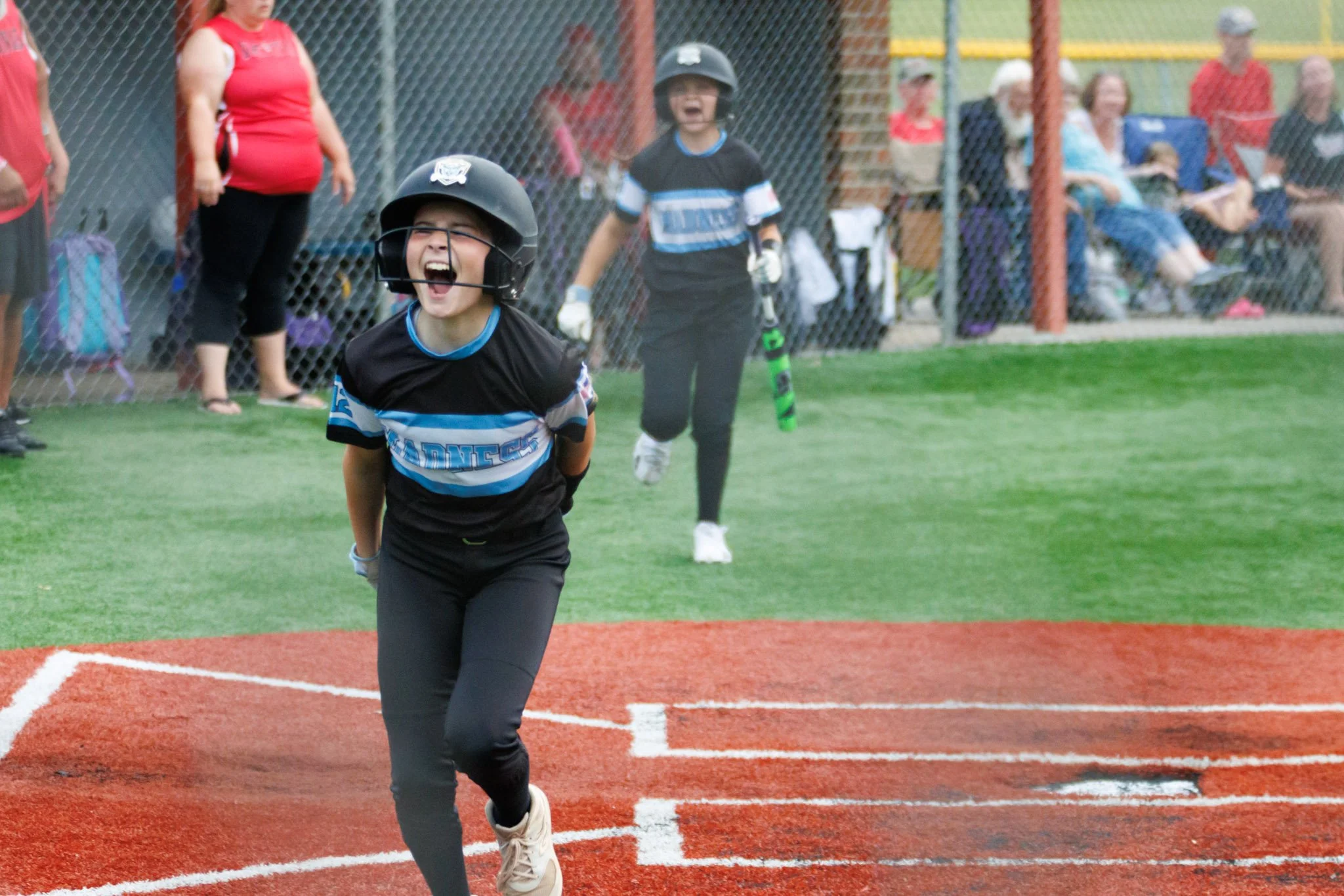 A young girl in a baseball uniform running on home plate during a softball game, with a girl in similar uniform and helmet following behind. Spectators are in the background behind a fence.