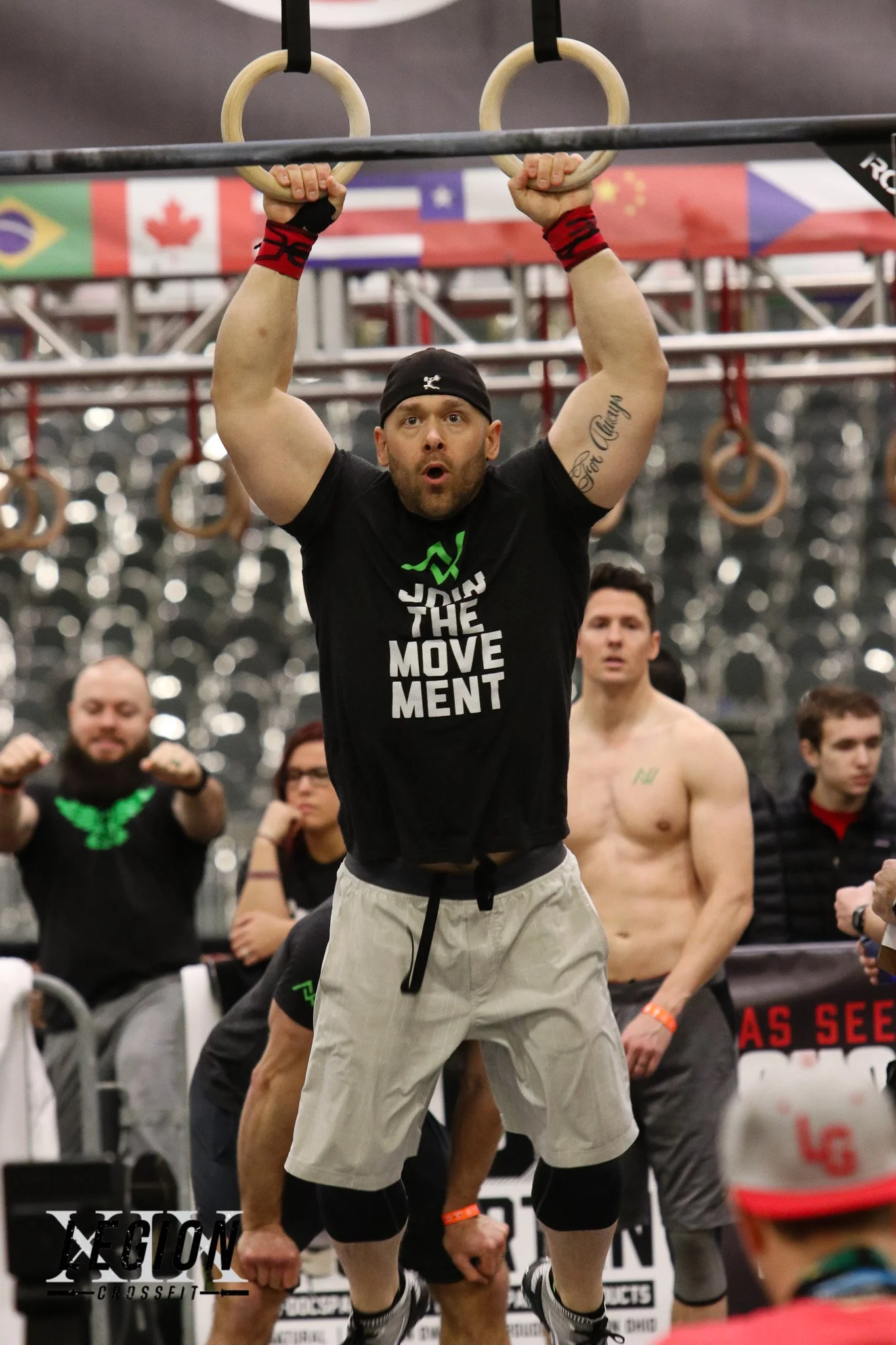 A muscular man with a black headband and wristbands doing pull-ups on gymnastic rings, surrounded by other athletes in a gym, with flags hanging from the ceiling.