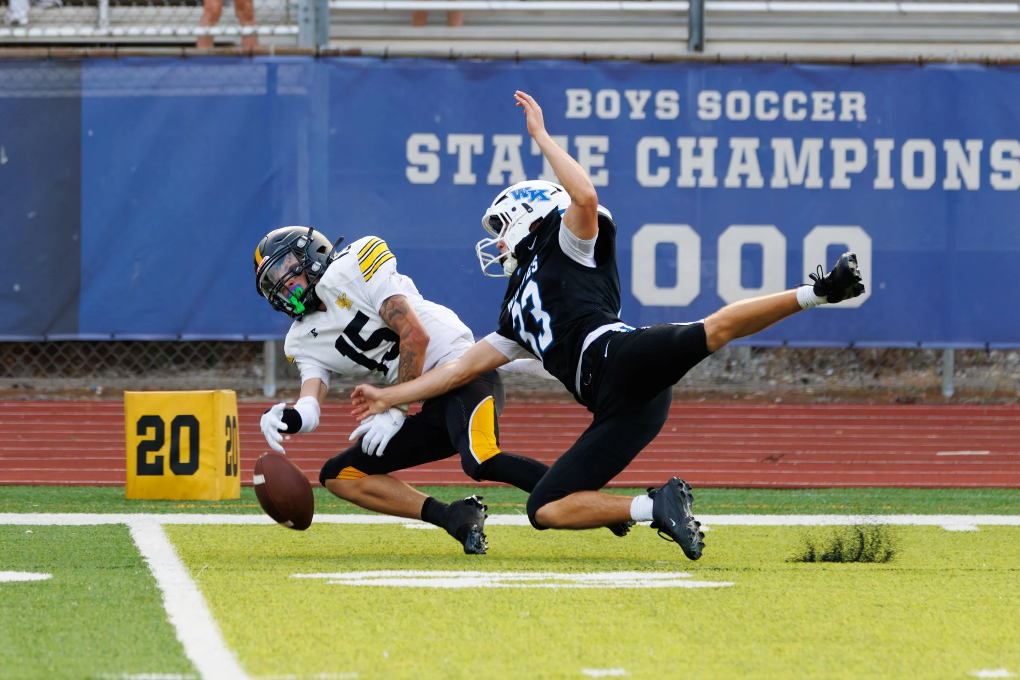 Two football players in helmets and uniforms collide on a field near the end zone, one attempting to catch the ball while the other tries to tackle him. A sign behind them reads 'Boys Soccer State Champions' and the number 20.