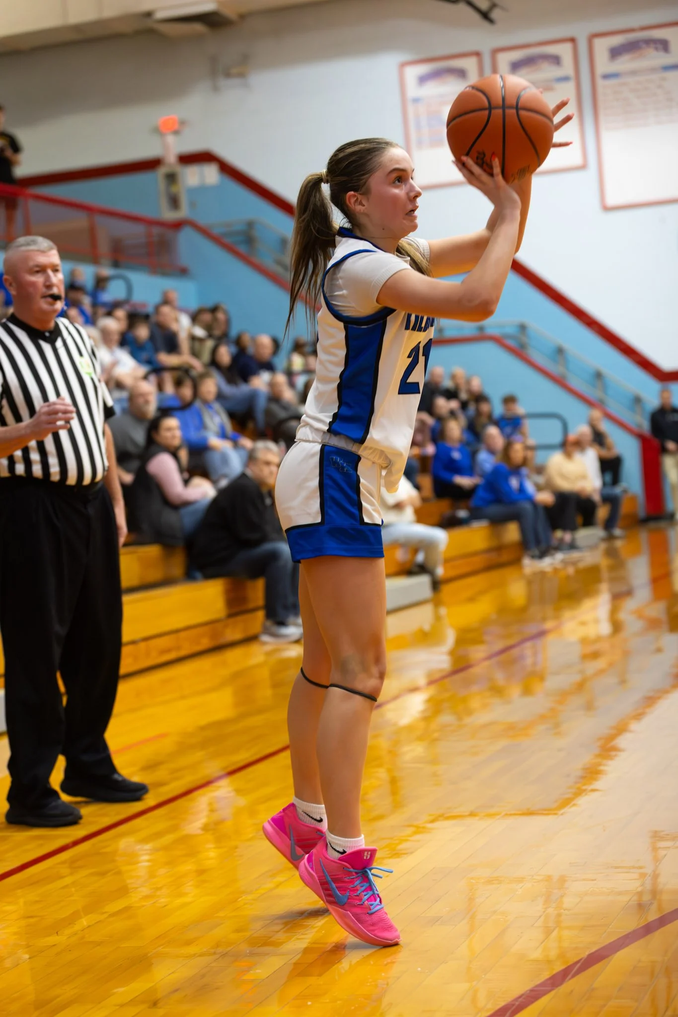 A female basketball player in a white and blue uniform shooting a free throw in a gymnasium, with spectators seated on bleachers in the background.