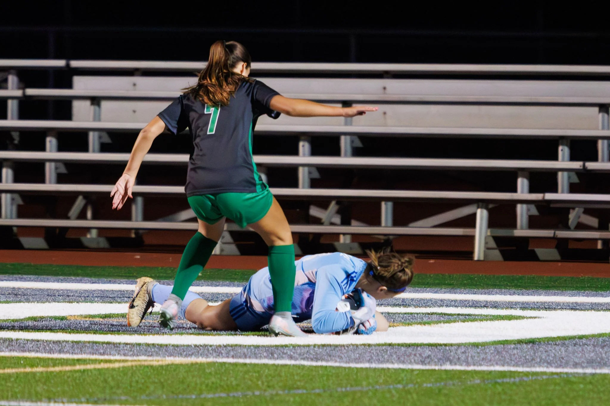 A female soccer player in a green and black uniform is attempting to score a goal near the sideline, while another female goalkeeper in a light blue uniform lies on the ground holding the ball.