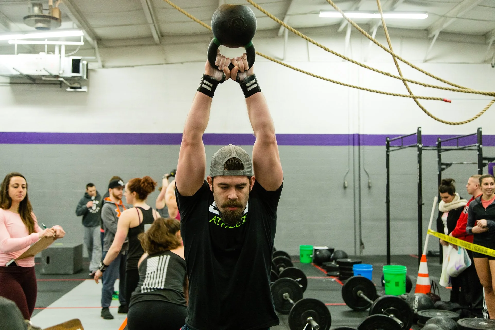 At a Crossfit competition, a man with a beard and gray backward cap performing a kettlebell lift in a gym. Several people are in the background, some with clipboards and others near gym equipment, with a yellow caution tape on the right side.