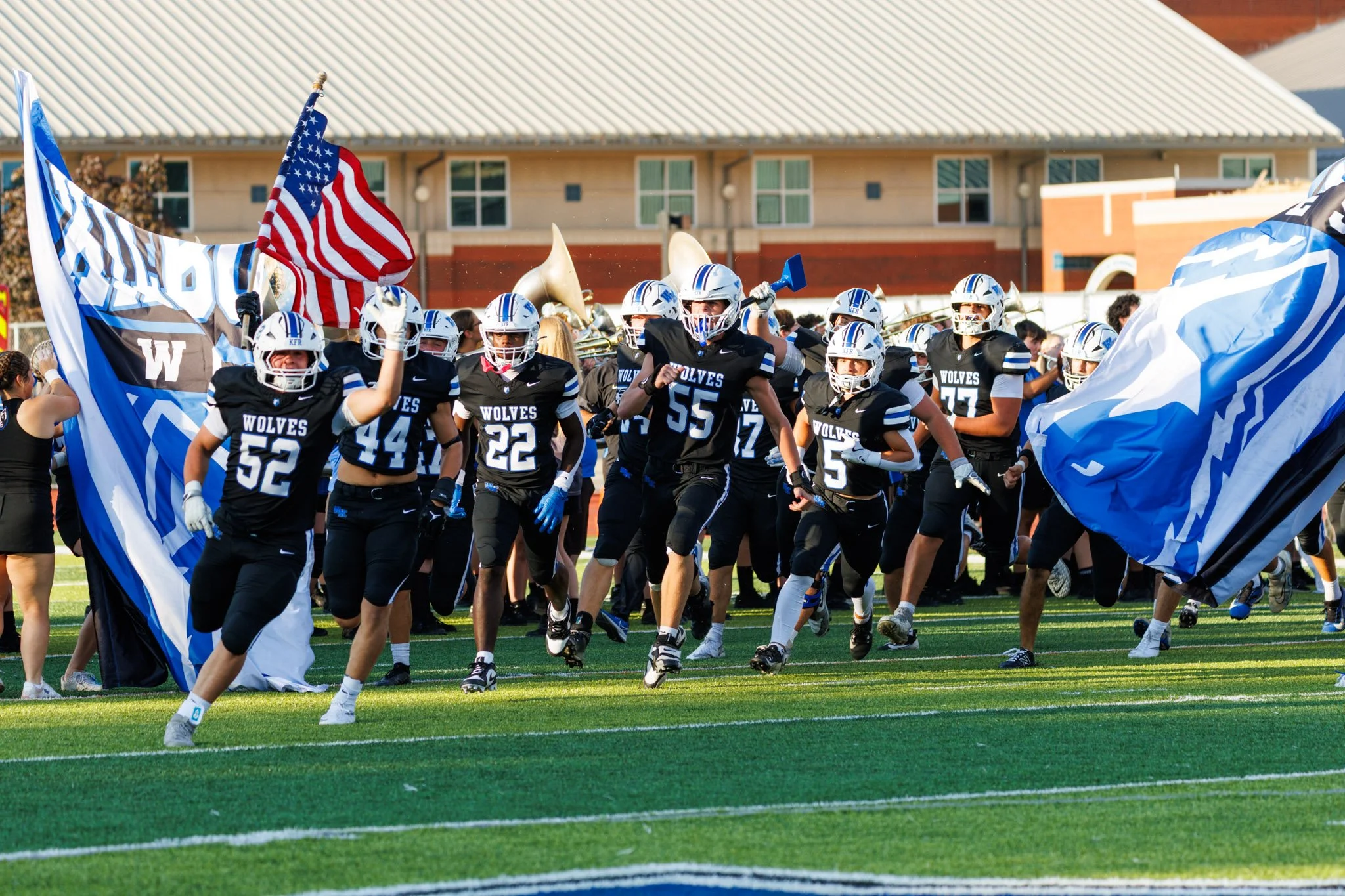 Kilbourne Wolves entering the field through a flag tunnel, wearing black uniforms with blue and white accents, some players holding flags and American flags, on a green football field in daylight.