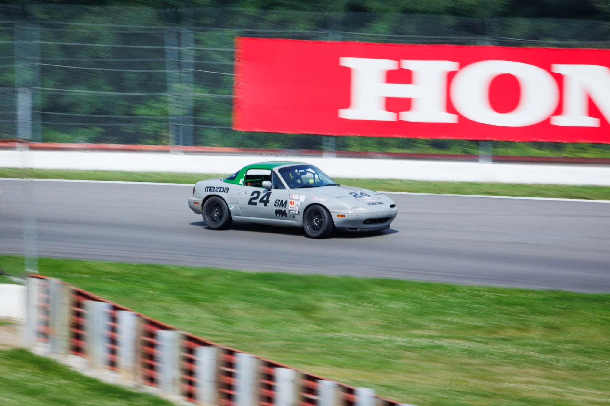 A silver race car with black wheels, number 24, speeding on a race track with a large red Honda advertisement in the background.