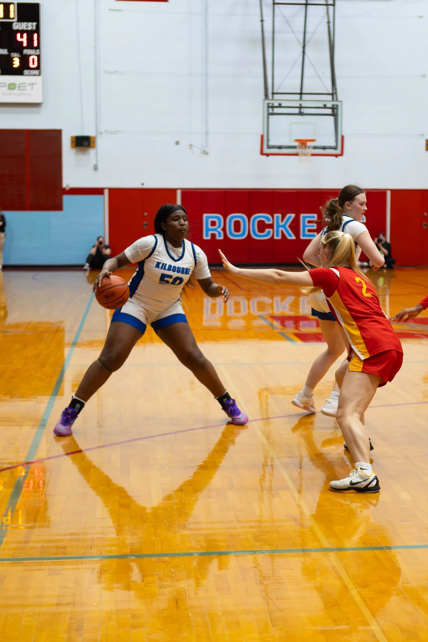 A women's basketball game in progress on a gym court with players from Kilbourne and another team. A Kilbourne player with a number 50 jersey is dribbling while defending against a player in a red and yellow uniform extending her arm. The gym has a s
