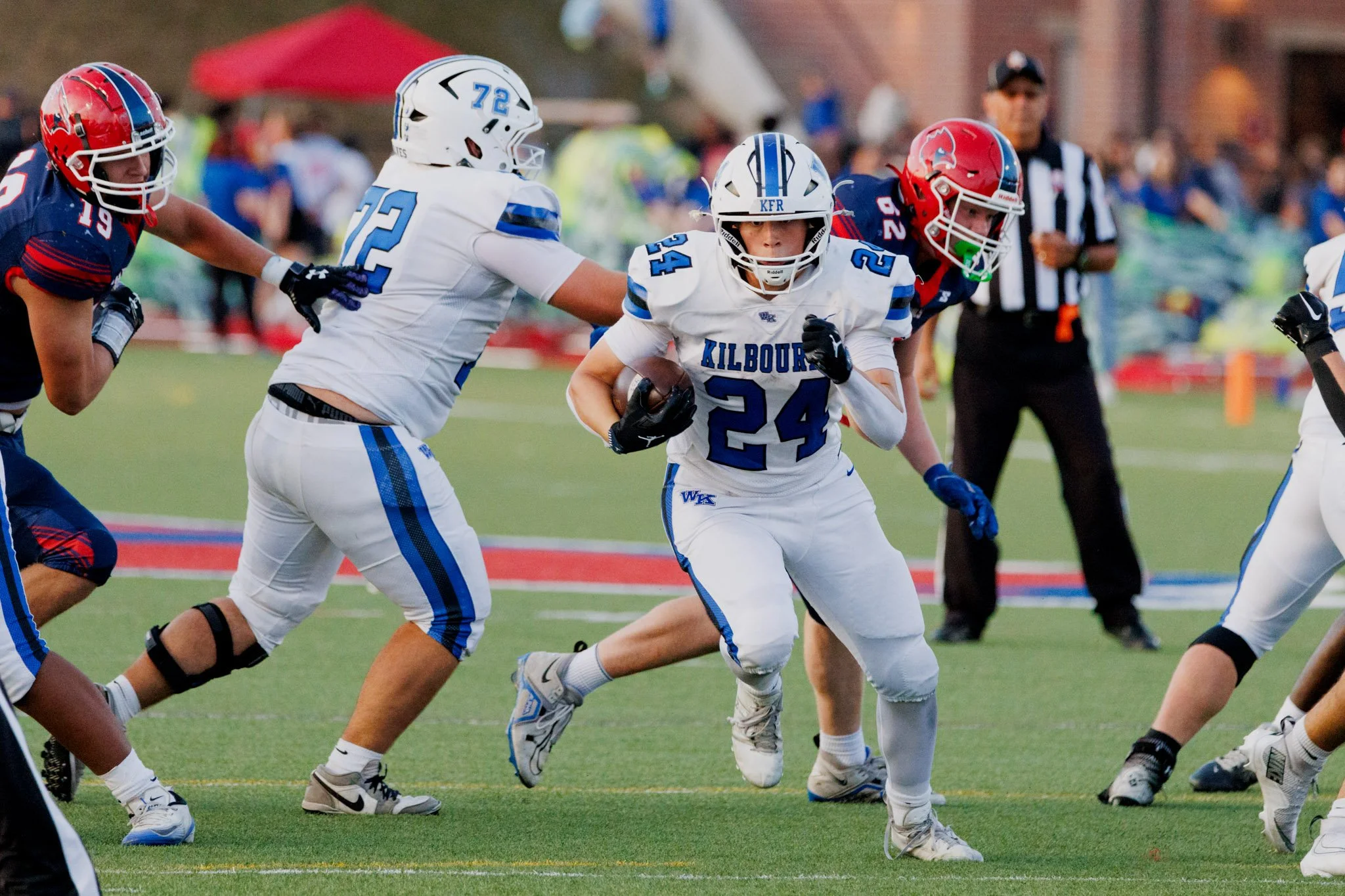 A group of football players in white and blue uniforms playing against players in navy blue and red uniforms on a football field with a referee in the background.
