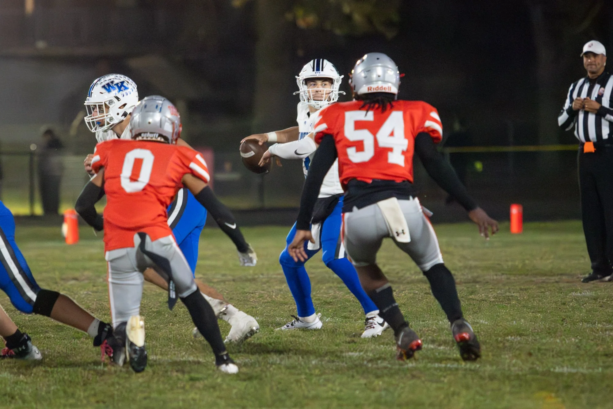 Football game with players in red and blue uniforms; the quarterback in blue prepares to throw, surrounded by opponents and defenders on a grassy field.