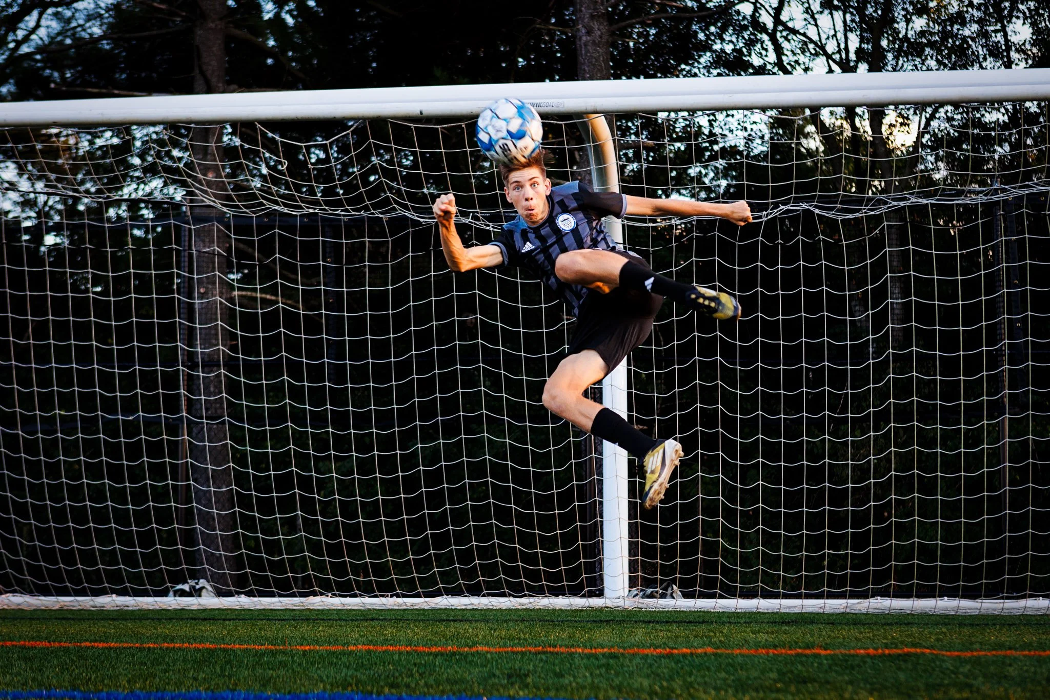 Soccer player in a navy and black striped uniform mid-air kicking a soccer ball during a game on a field with a goal net and trees in the background.