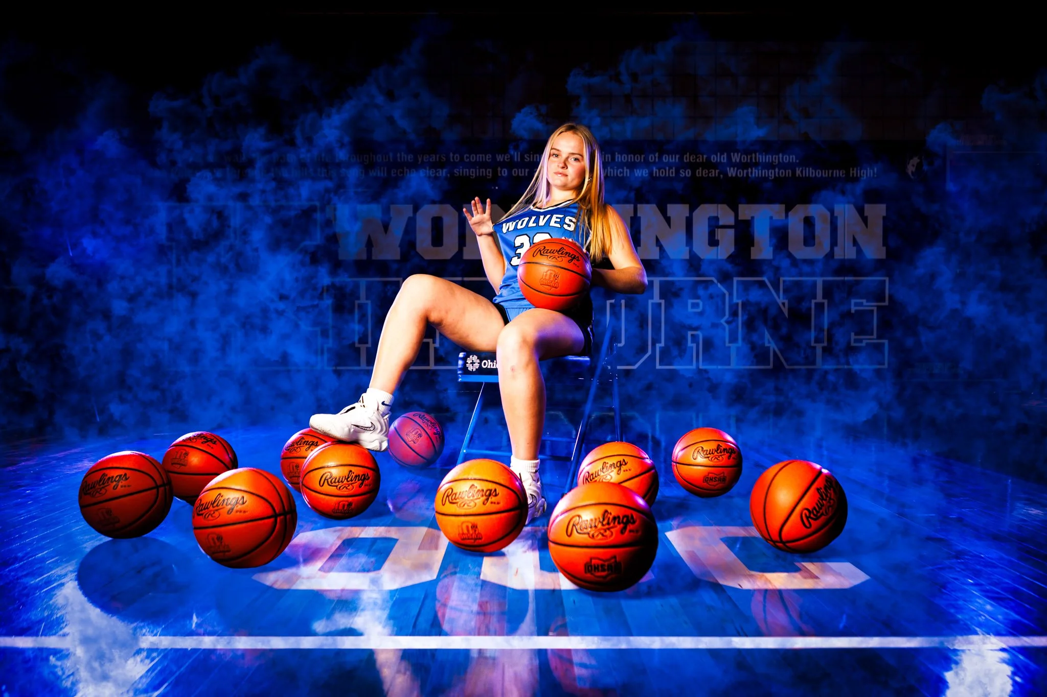 A young female basketball player in a blue jersey with 'Wolves' and the number 23, sitting on a chair in a gymnasium surrounded by basketballs, holding a basketball in her lap, with some basketballs on the floor around her, against a dark background 