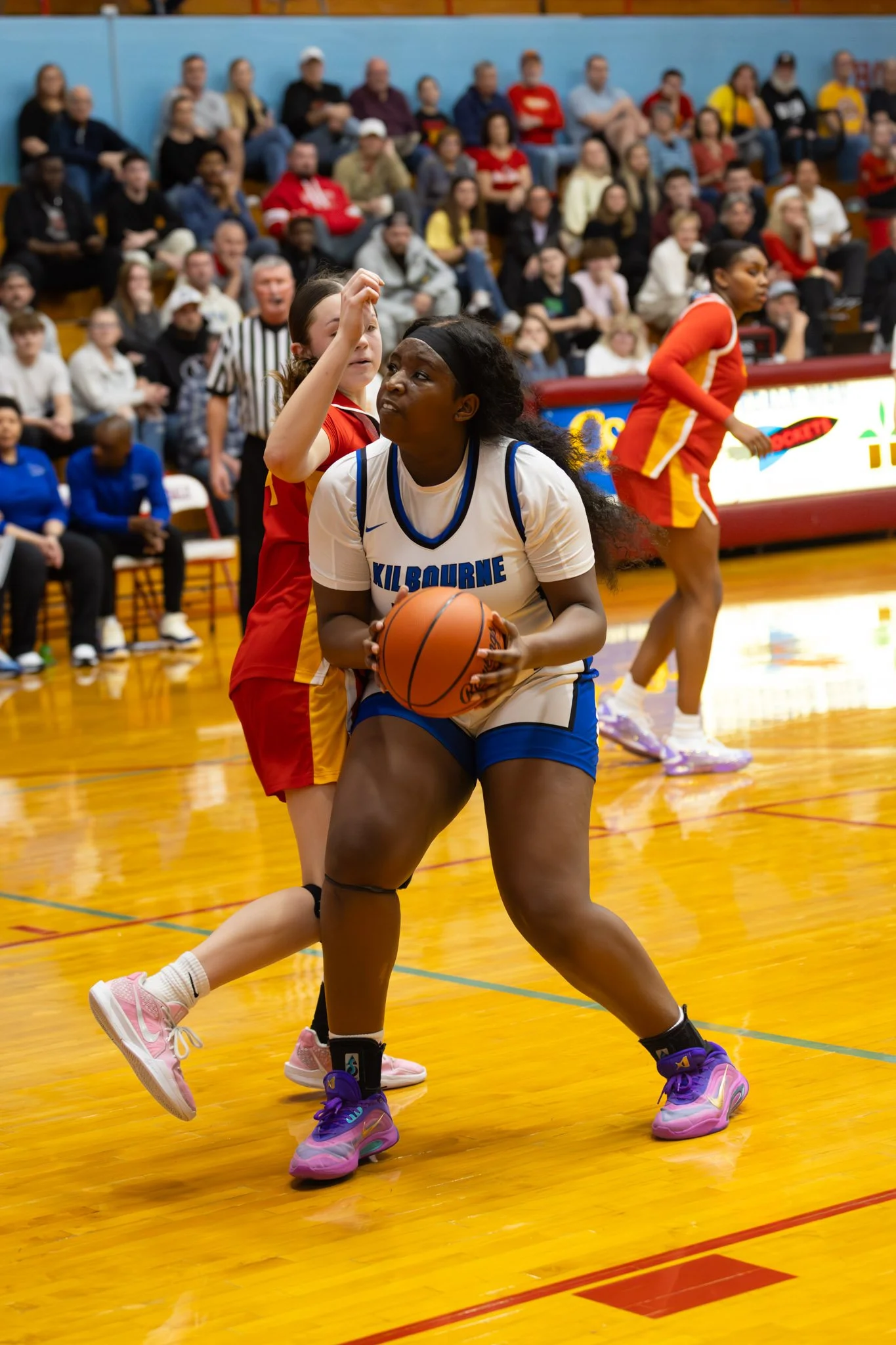 A female basketball player in a white and blue uniform holding a basketball, guarded closely by an opponent in a red and yellow uniform near the basket during a game, with an audience in the background.