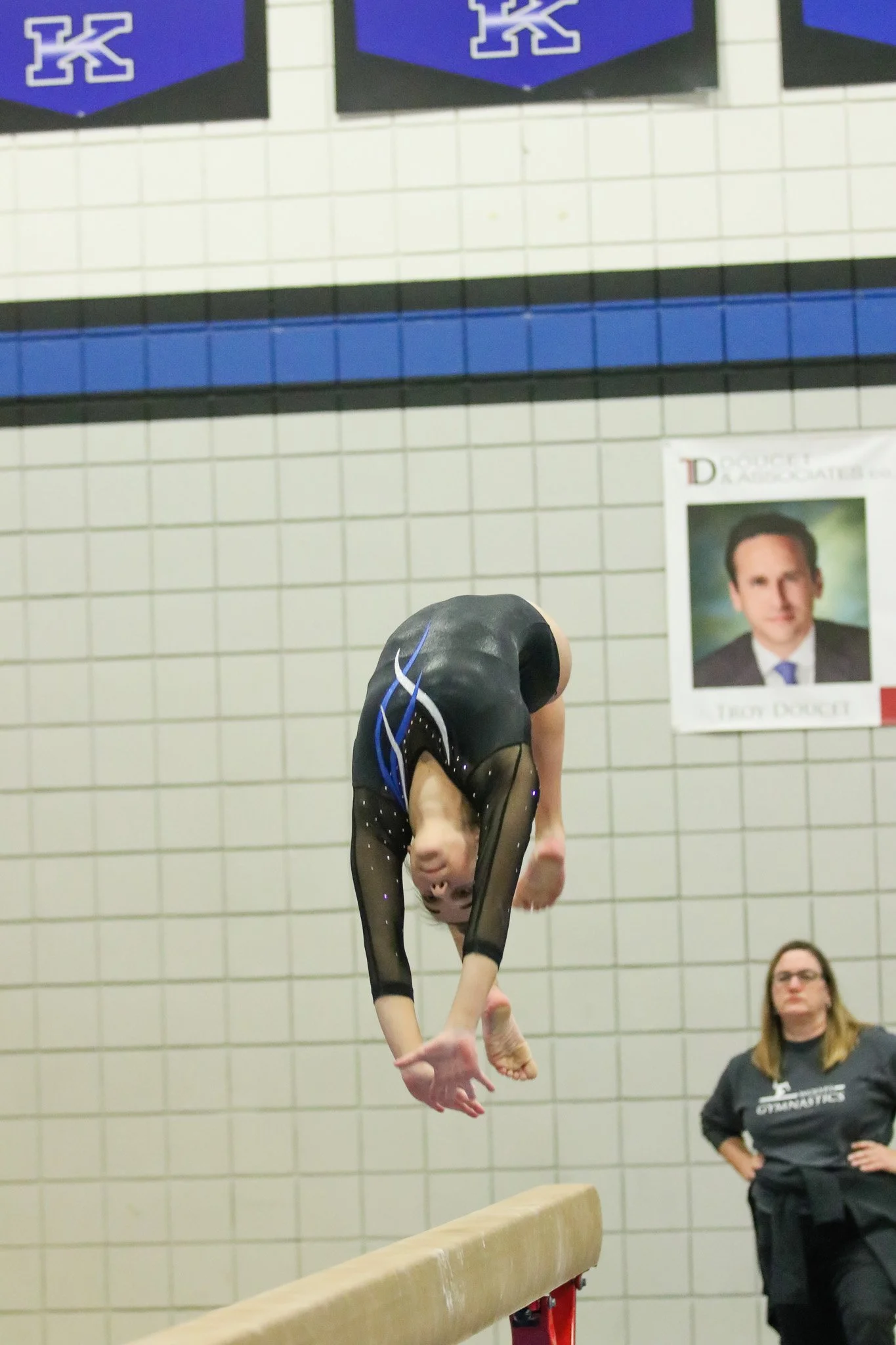 A female gymnast is performing a balance beam routine in a gymnasium. She is upside down with her arms extended, wearing a black leotard with blue accents and mesh sleeves. 