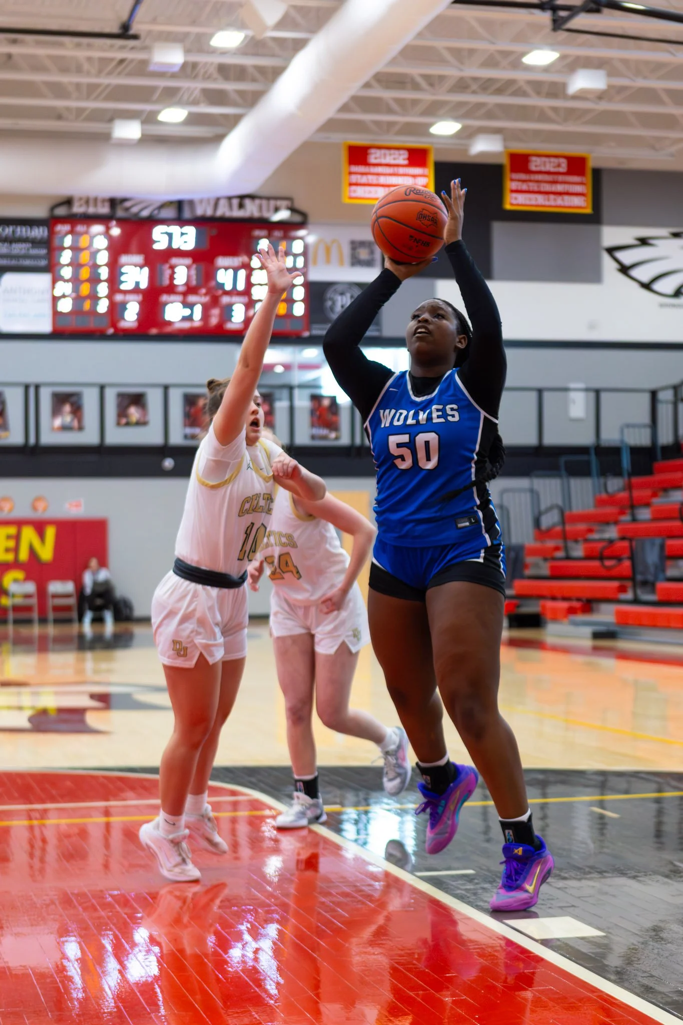 A basketball player in a blue jersey with number 50 and 'WOLVES' written on it, jumping to shoot the ball in a game. Two players in white jerseys with gold accents are defending. The game is in a gymnasium with a red and black scoreboard and empty bl