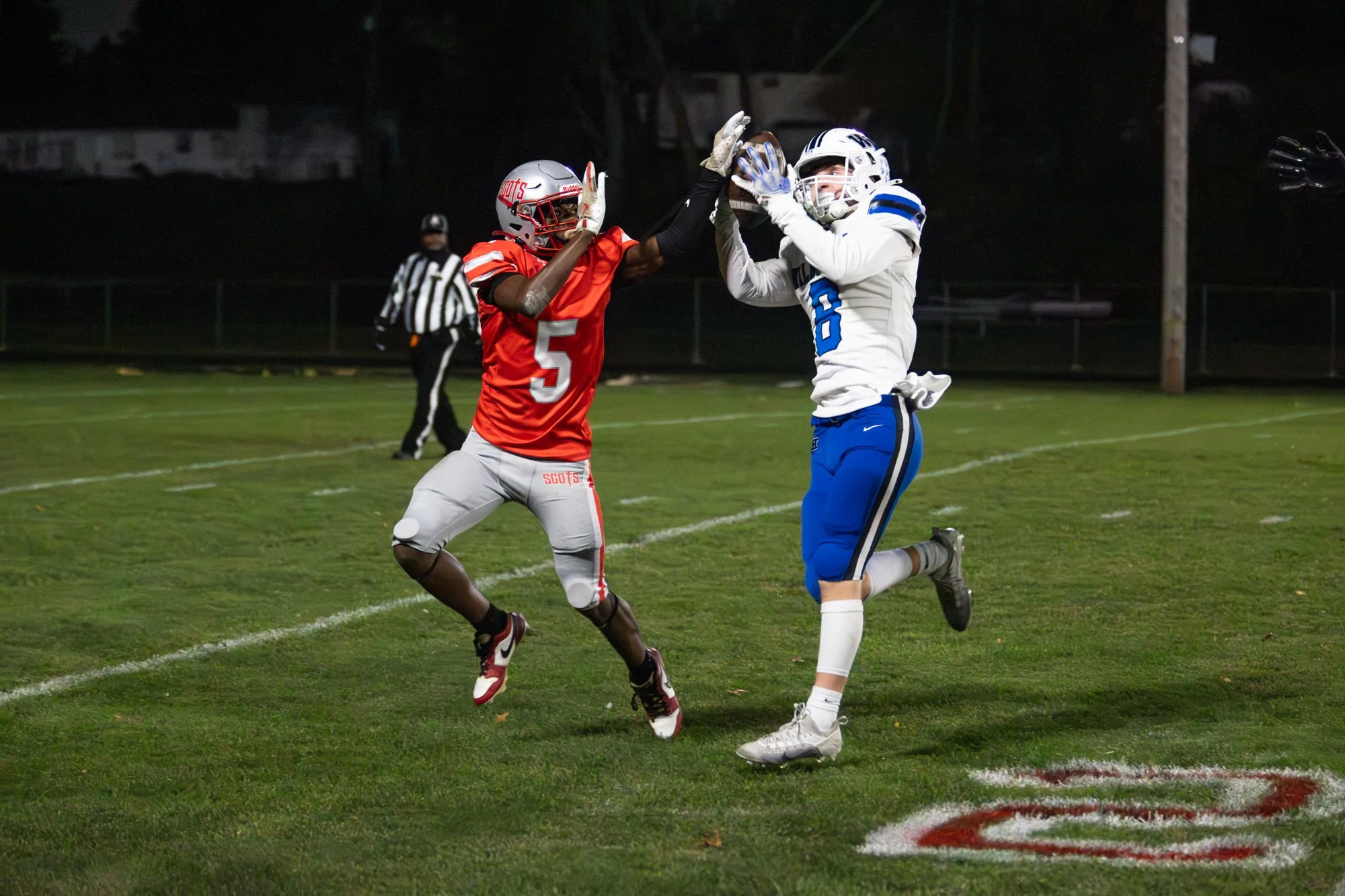 Two football players in mid-air on a field at night during a game, with an referee in the background.