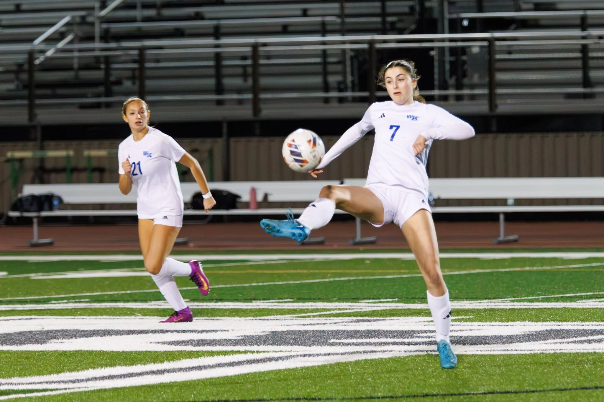 Two female soccer players in white uniforms playing on a field at night. One player with jersey number 7 is kicking a soccer ball, while the other player with jersey number 21 is running nearby.