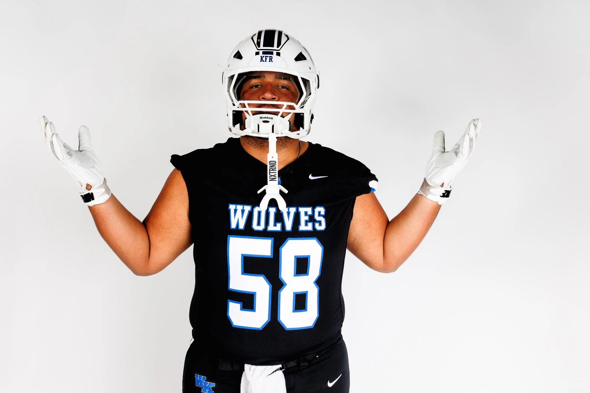 Media Day shoot.  Football player wearing a white helmet with black and blue accents, a black jersey with blue and white lettering that reads 'WOLVES' and the number 58, standing with arms raised against a plain white background.