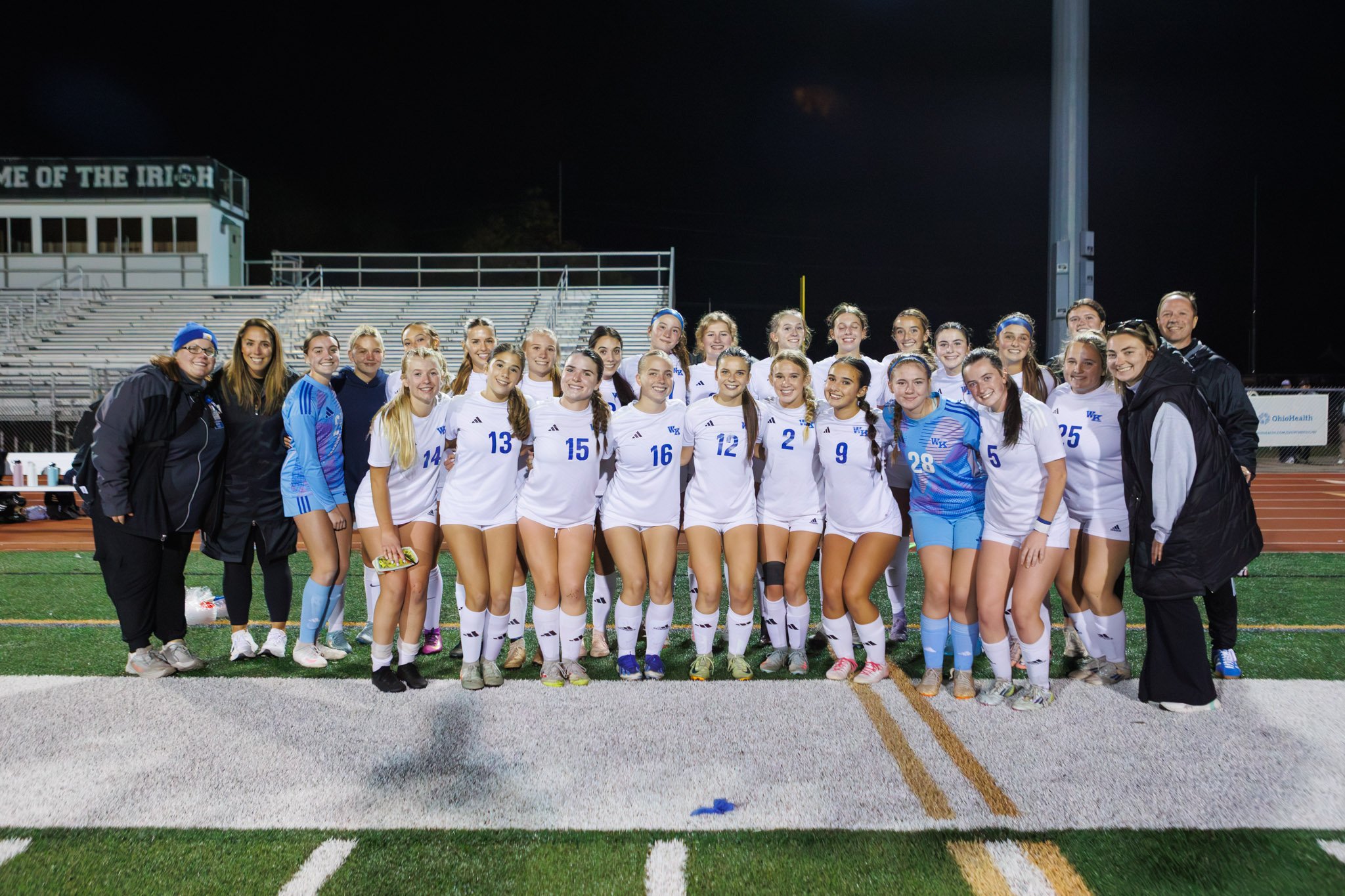 A women's soccer team poses for a group photo on a field at night, wearing white jerseys with blue accents, alongside their coaches and staff.