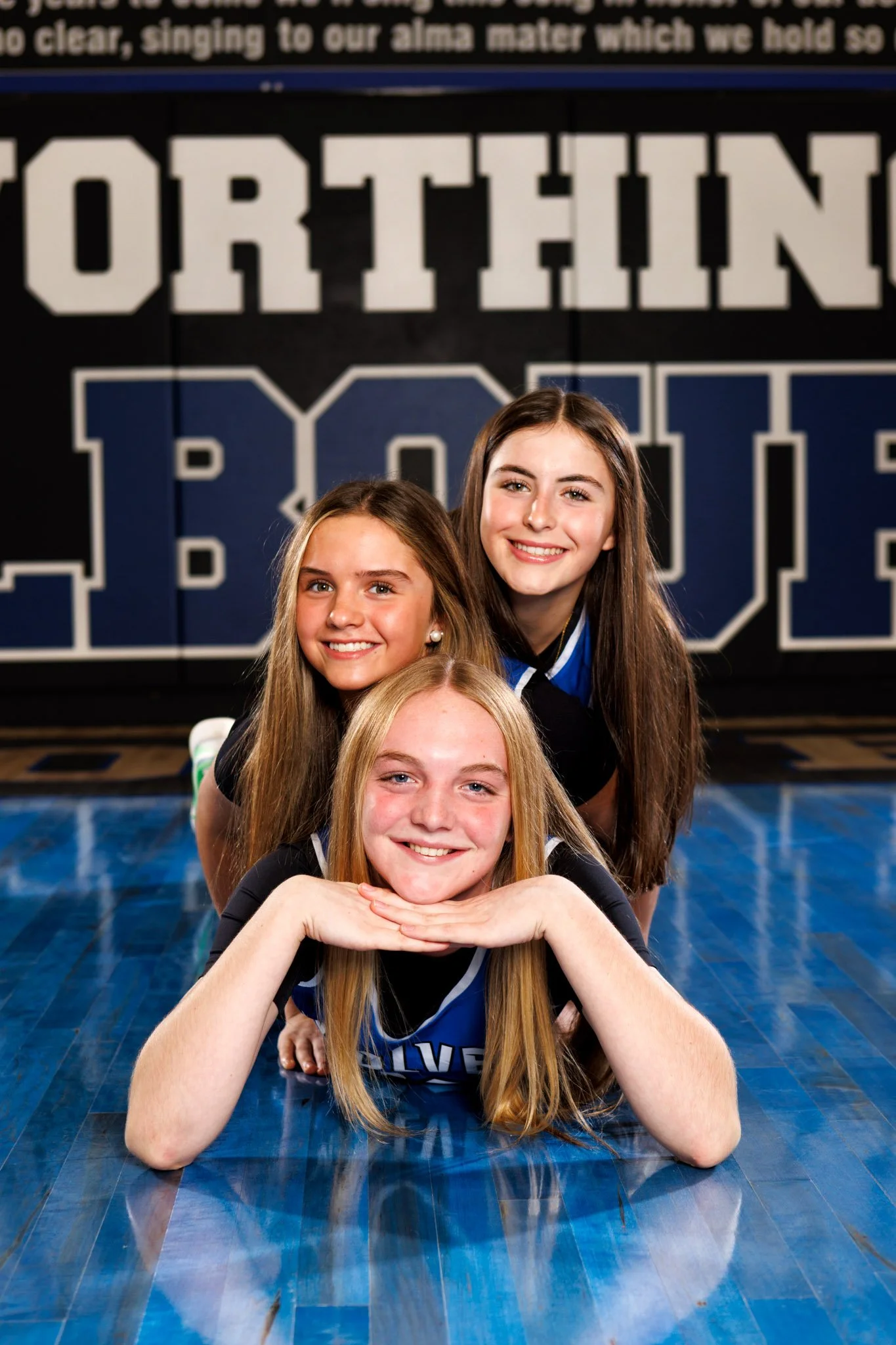 Shots from a media day shoot.  Three young women in sports uniforms, all smiling, lying on a gymnasium floor with a large sports-themed sign in the background.
