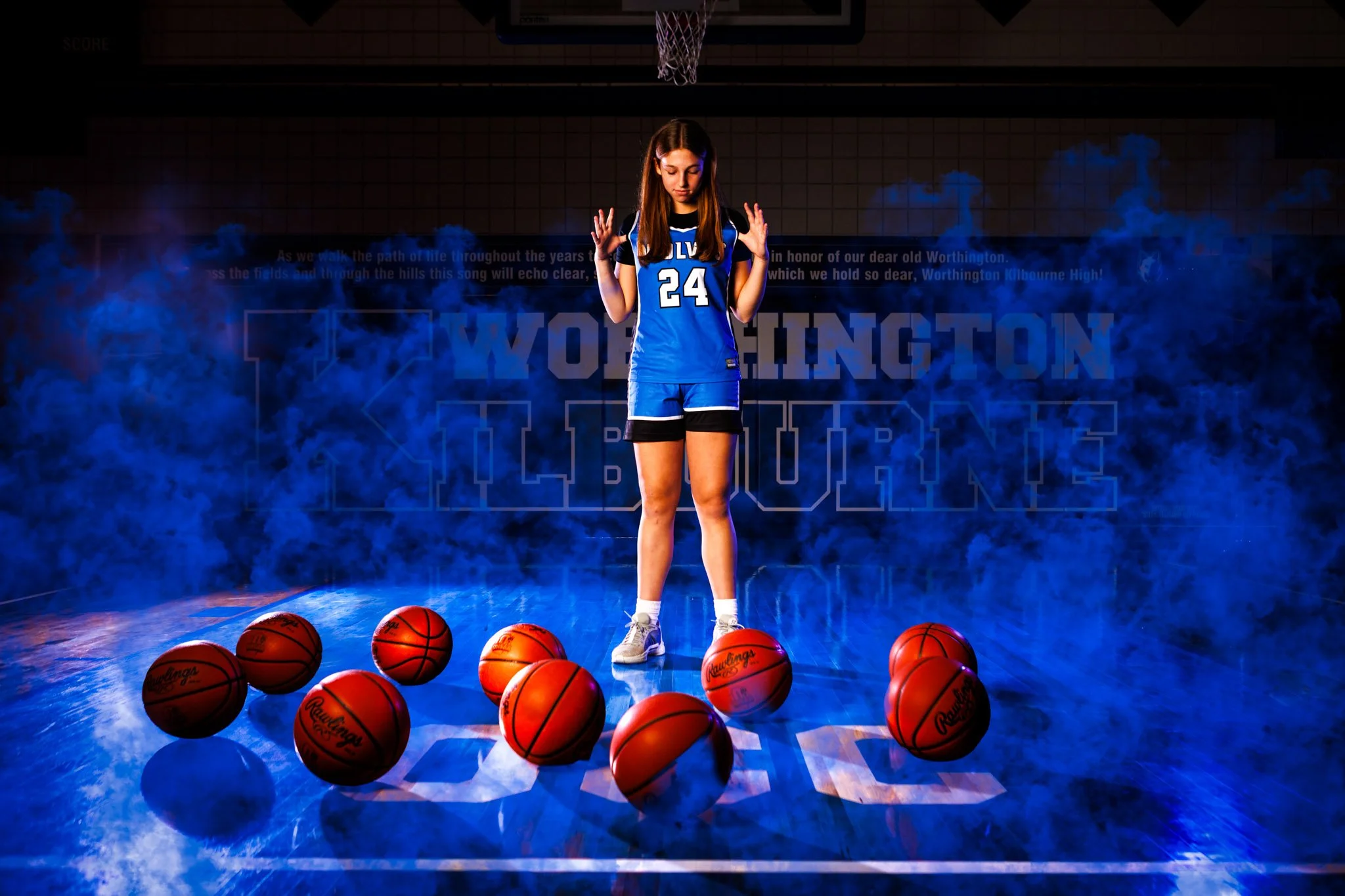 Media day shoot.  A female basketball player in a blue jersey with the number 24, standing on a basketball court surrounded by nine basketballs. The background features a dark gymnasium with blue lighting and a haze of smoke, with the words ‘WORTHING