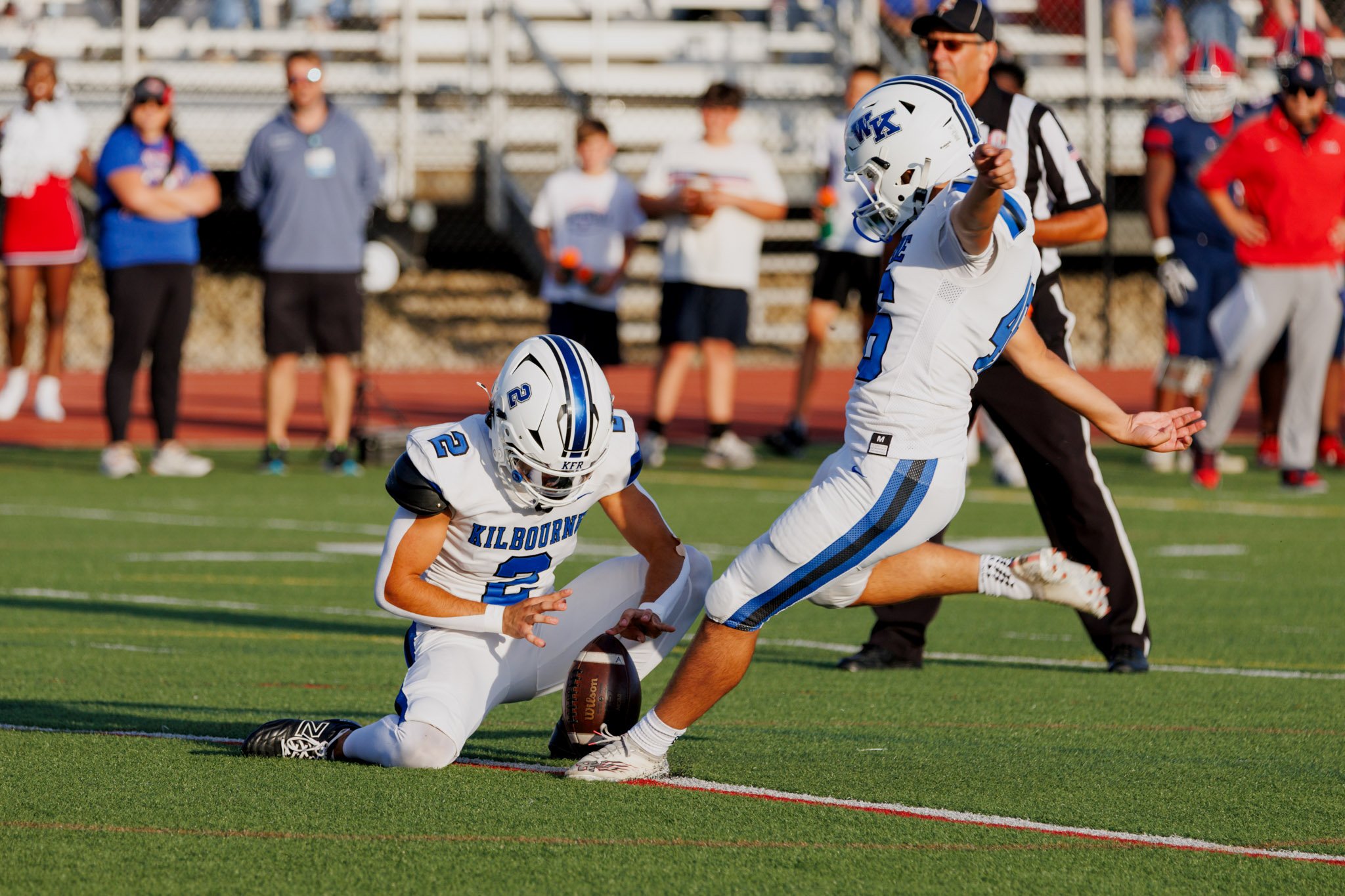 Two football players in white and blue uniforms from Kilbourne High School attempting an extra point kick.