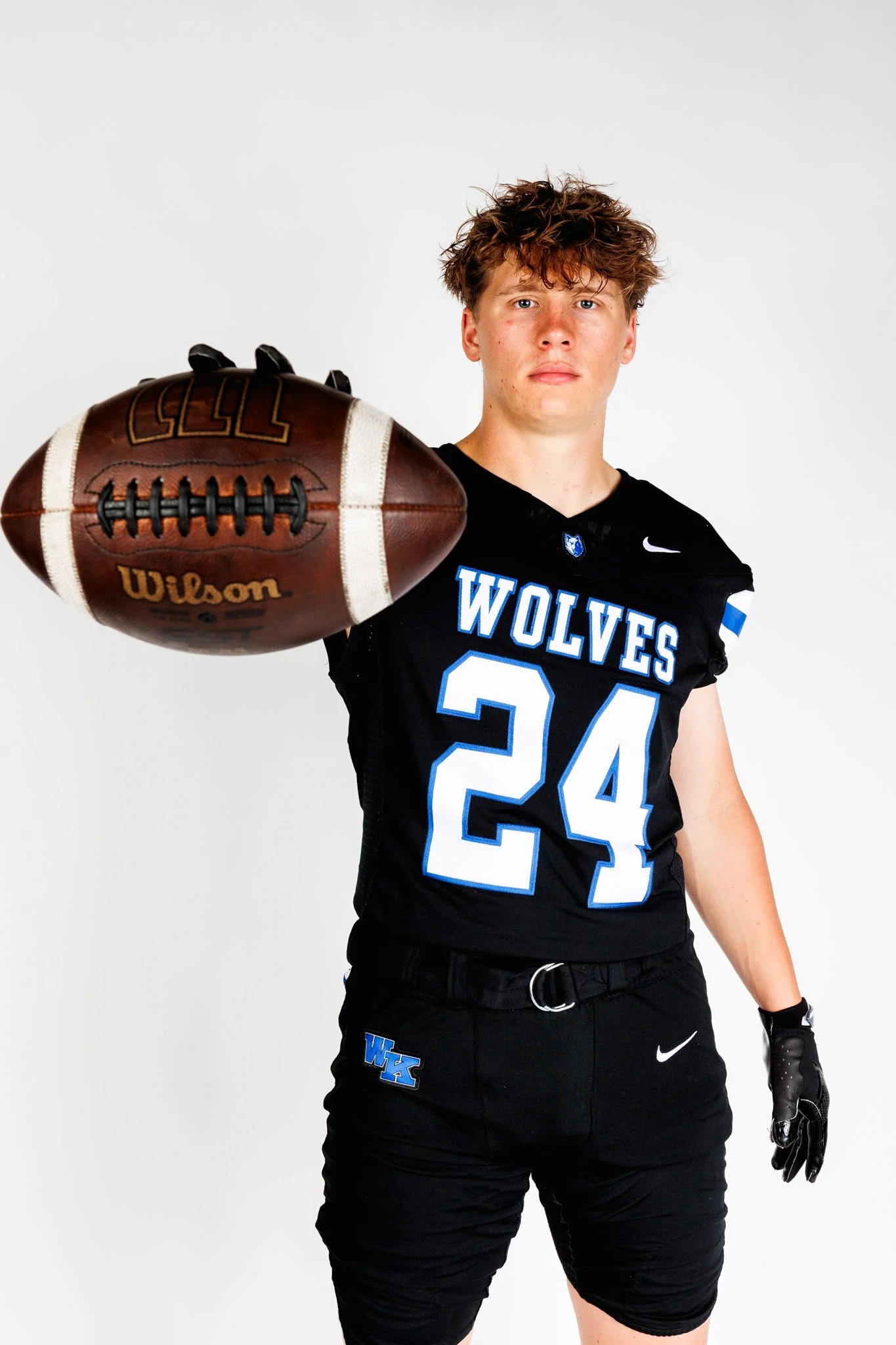 Shots from a media day shoot. A young male football player with curly hair, wearing a black team jersey with 'WOLVES' and the number 24, holding a football extended toward the camera, standing against a plain white background.
