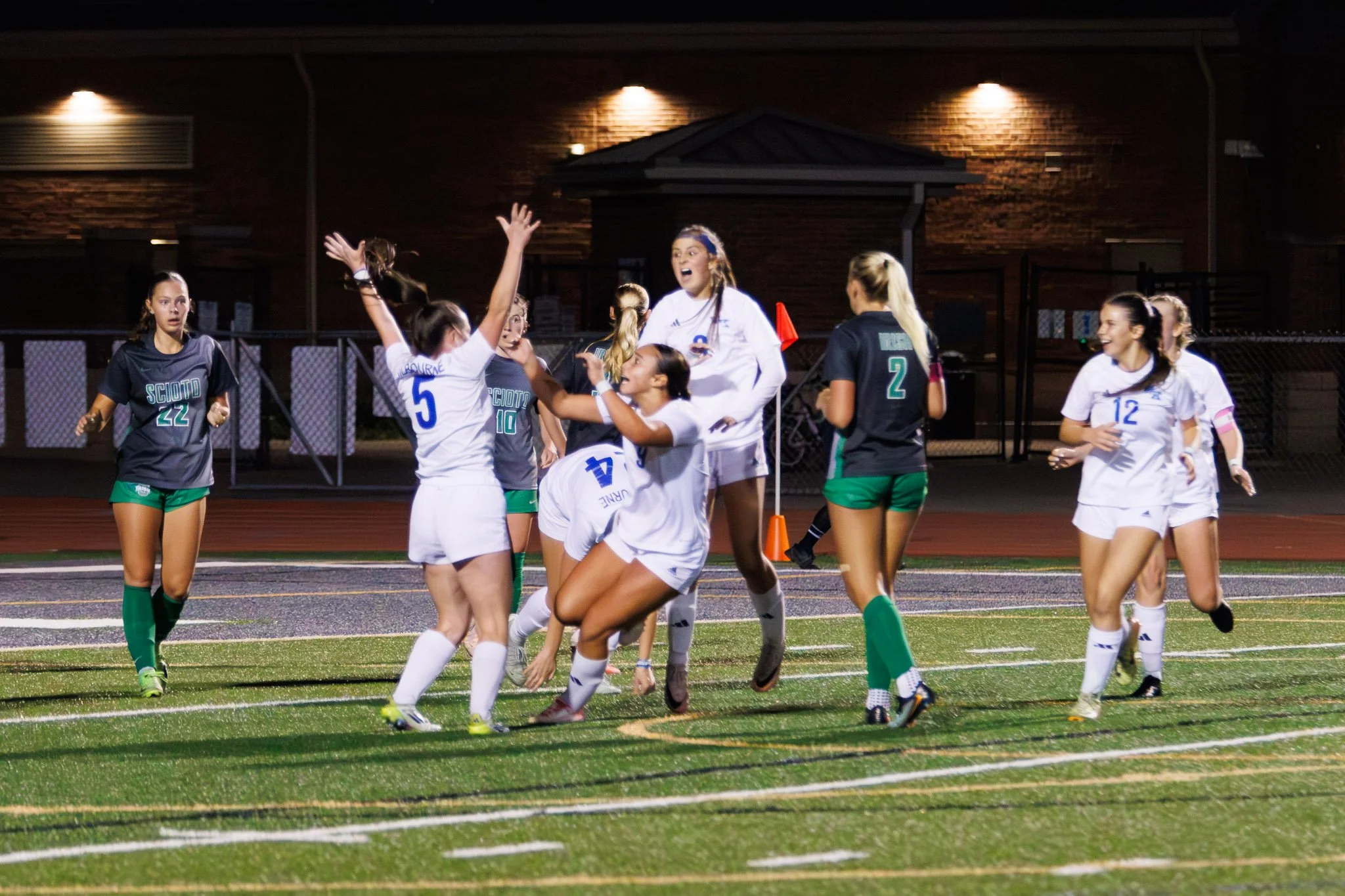 Women’s soccer team celebrating on the field after scoring a goal during a game at night.
