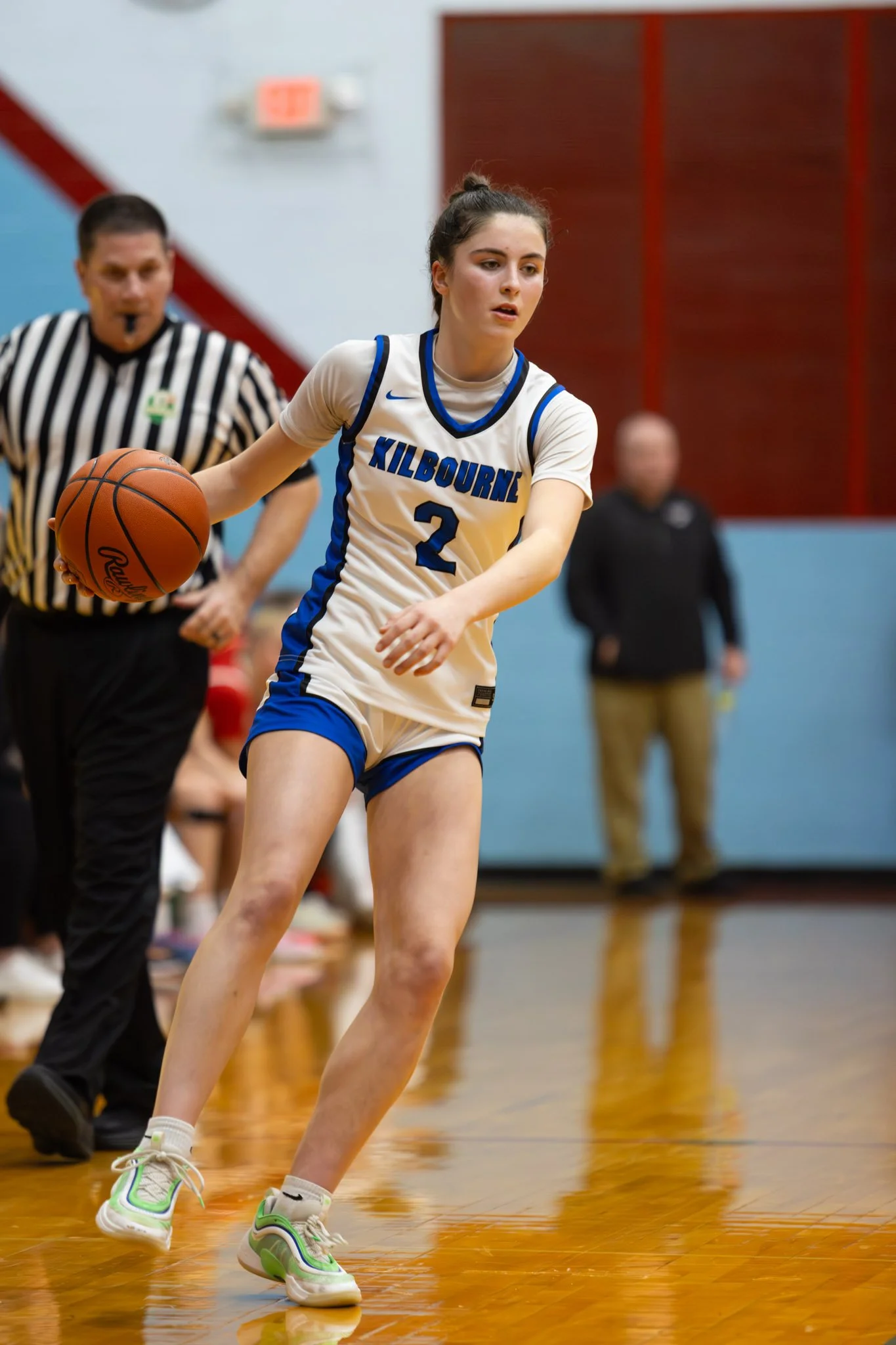 A young female basketball player in a white and blue uniform with the word 'KILBOURNE' and the number 2, dribbling a basketball during a game in a gymnasium.