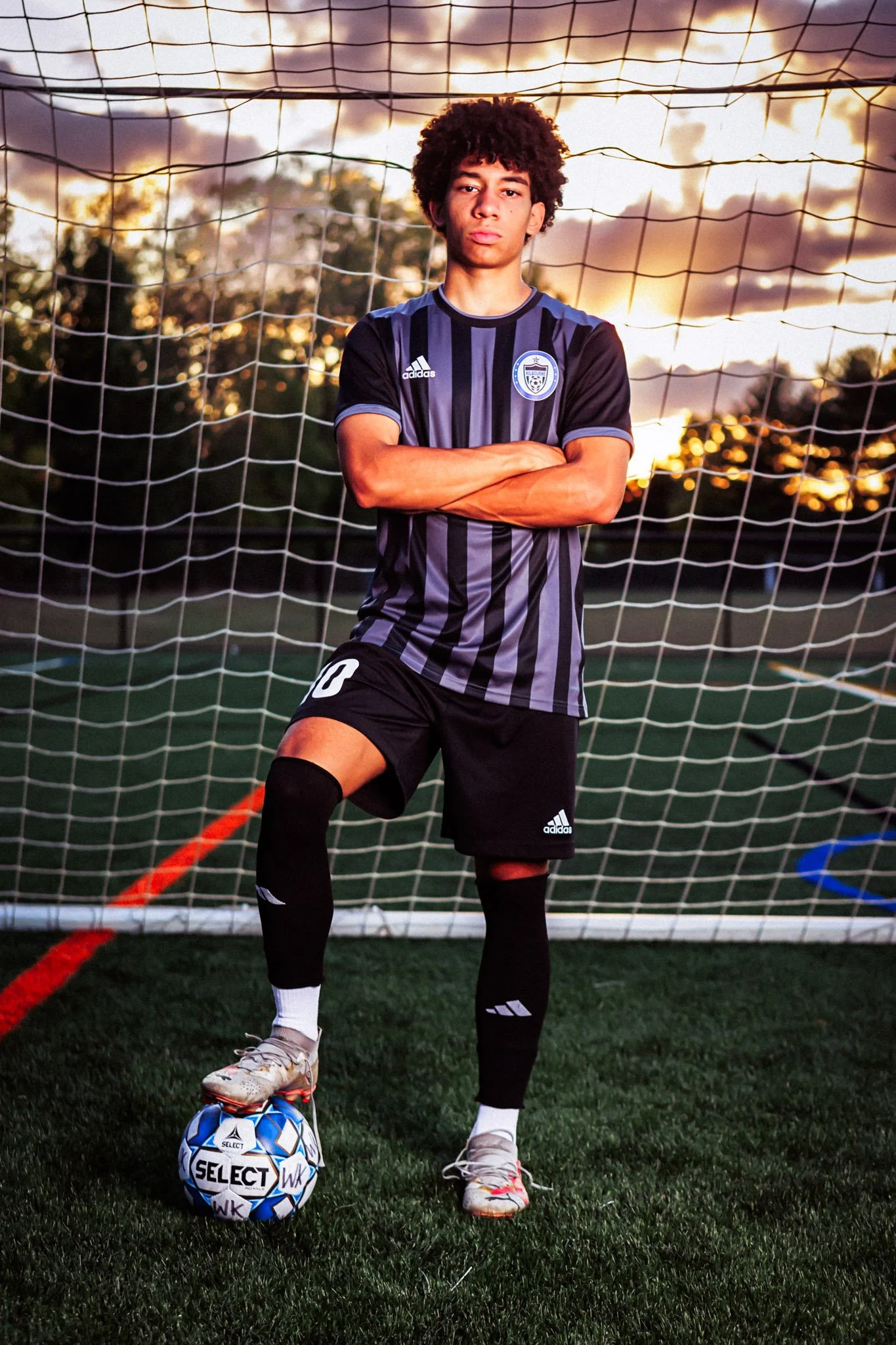 A young male soccer player standing on a soccer field in front of a goal net, with his arms crossed and one foot resting on a soccer ball, during sunset.