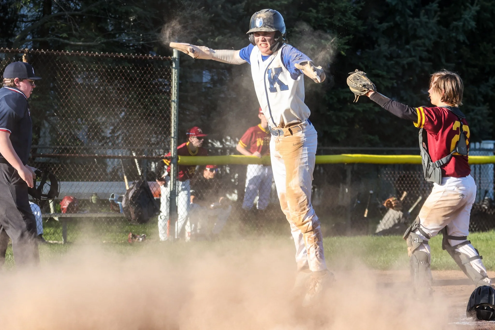 A baseball player wearing a blue and white uniform, with the letter 'K' on his jersey, is sliding into home plate as dust and dirt fly around. He is being tagged out by a catcher dressed in a red and black uniform with shin guards. An umpire stands n