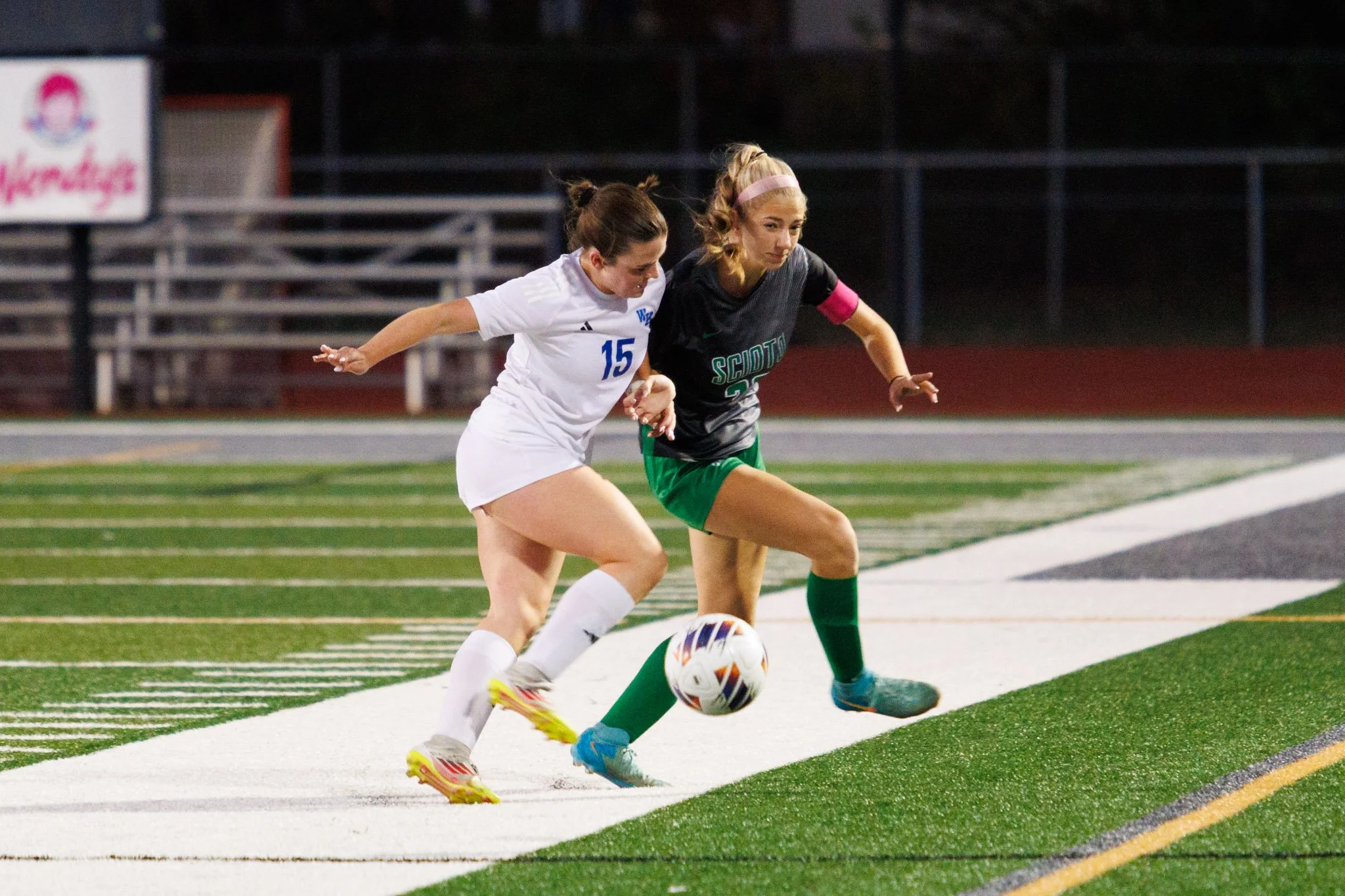 Two female soccer players competing for the ball near the sideline on a soccer field during evening game.