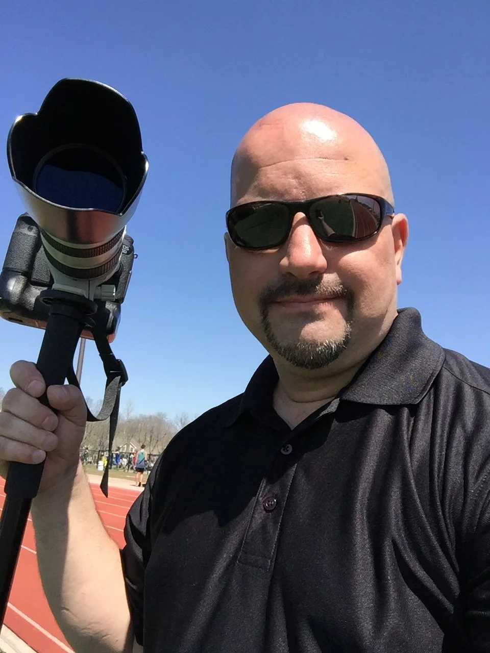 A man with sunglasses holding a professional camera with a large lens, standing outdoors on a track field on a sunny day.
