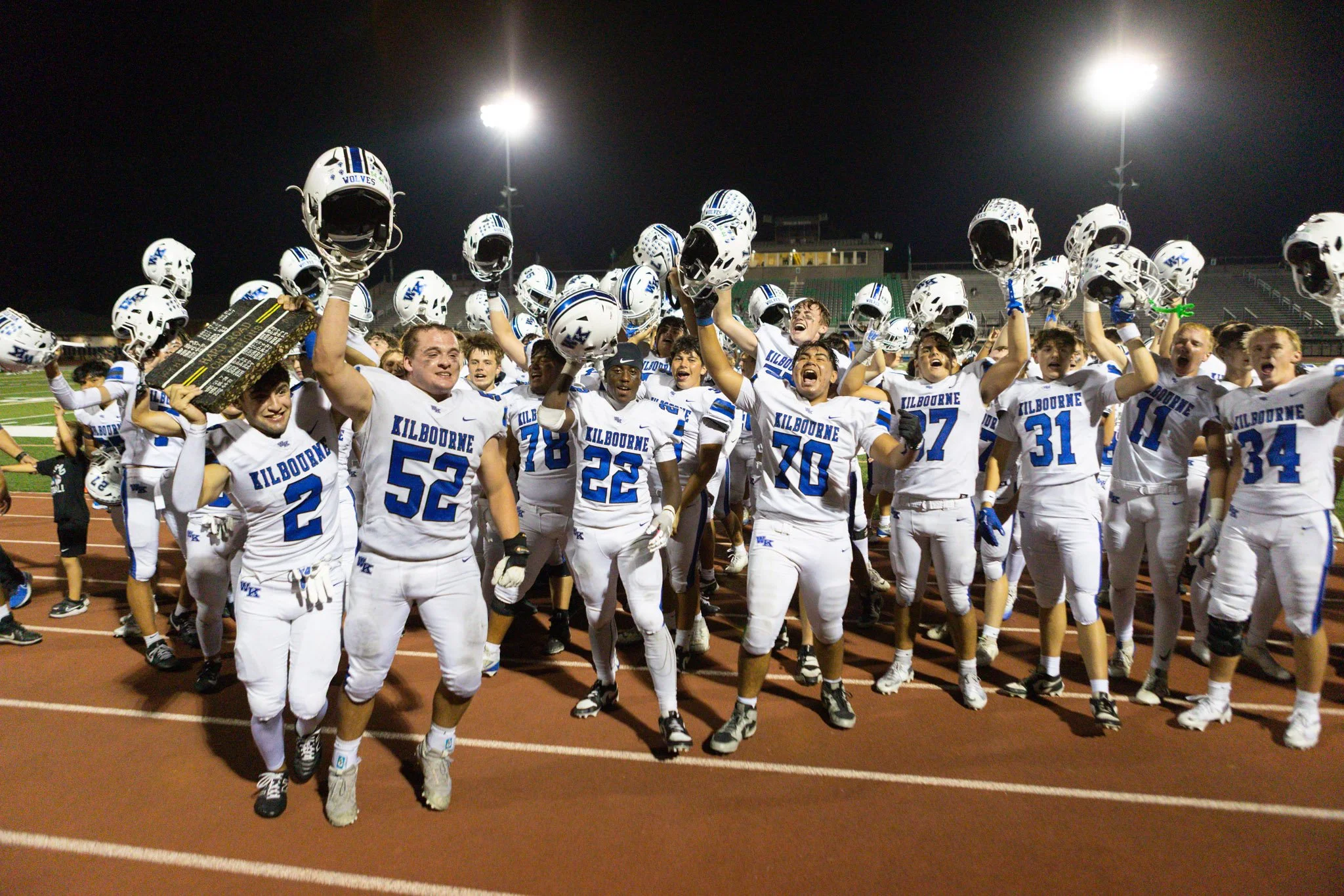 Football team from Kilbourne High School celebrating victory on field at night, lifting helmets 