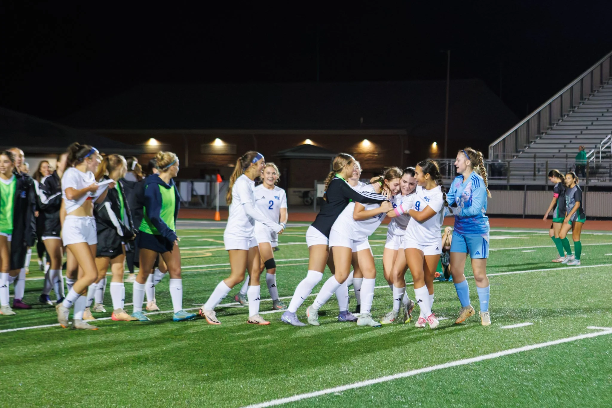 Girls soccer team celebrating on the field at night, with some players laughing and embracing each other, while teammates and coaches look on.
