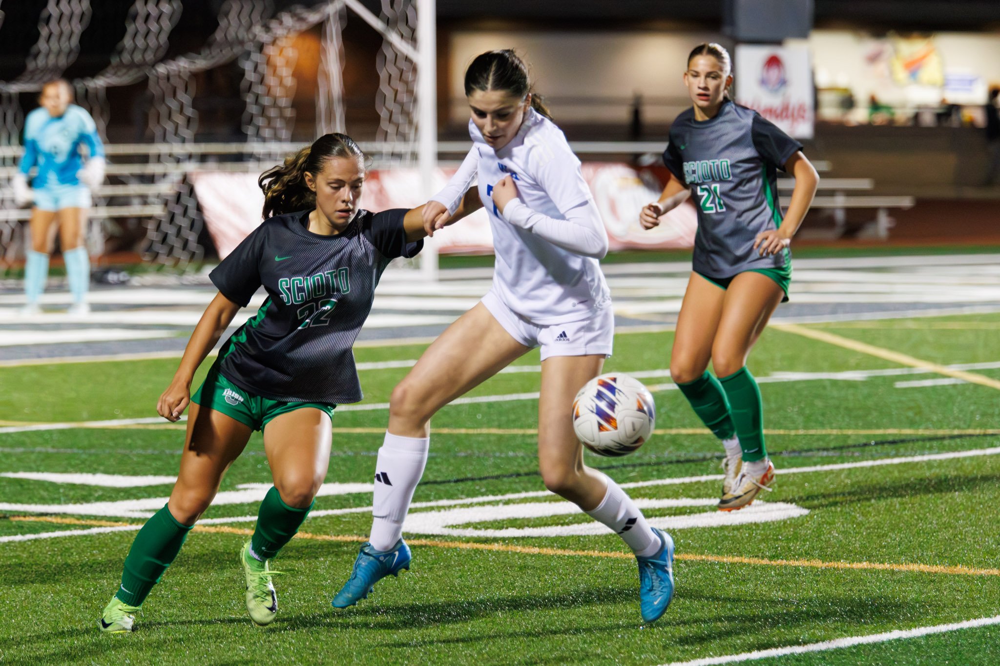 Three female soccer players competing for the ball during a match on a turf field, with players wearing black and white uniforms, and a goalkeeper in the background.