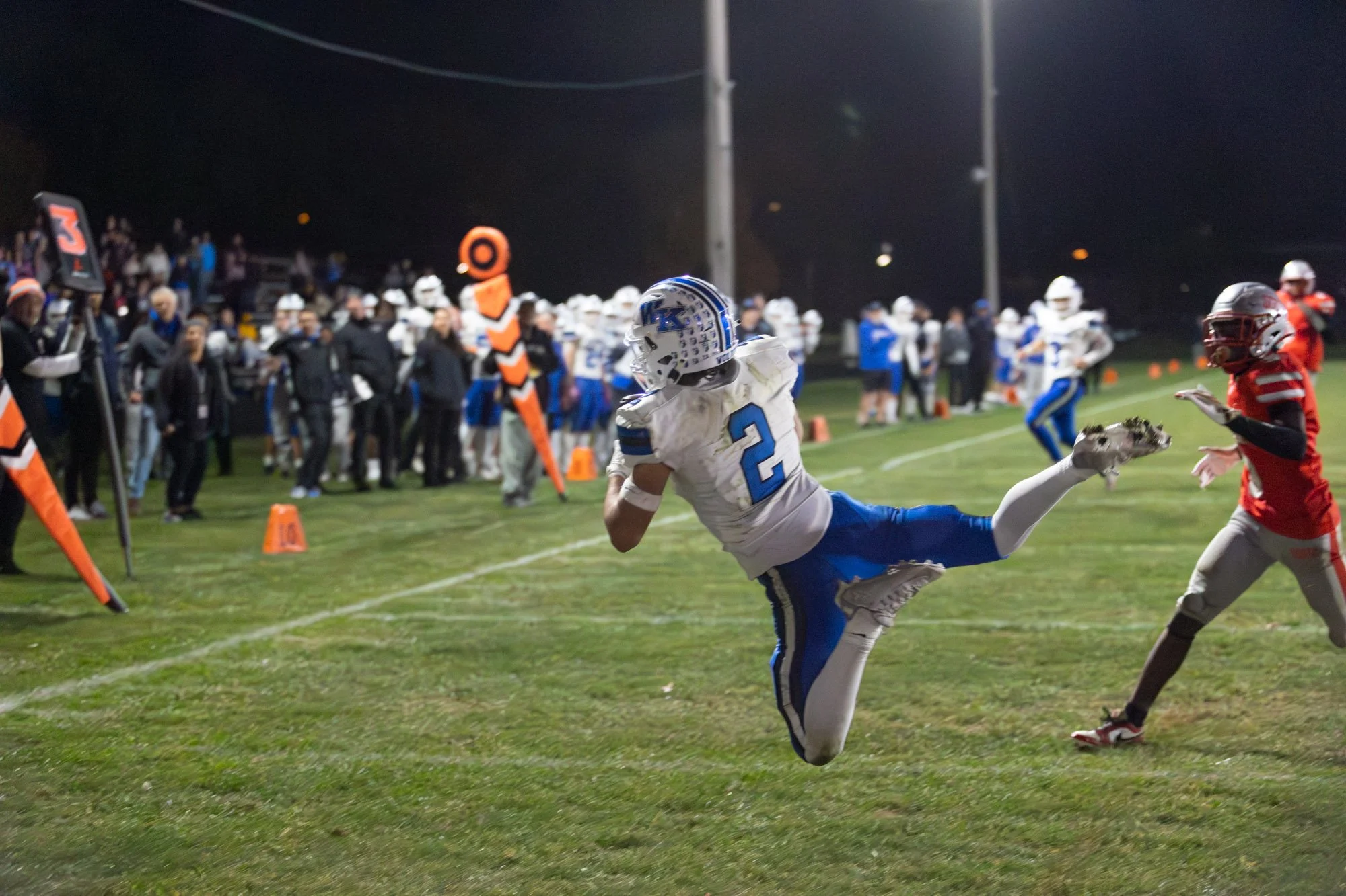 A football player wearing a white and blue uniform with the number 2 is catching a football while leaping during a game at night. The sideline with coaches, players, and officials is visible in the background.